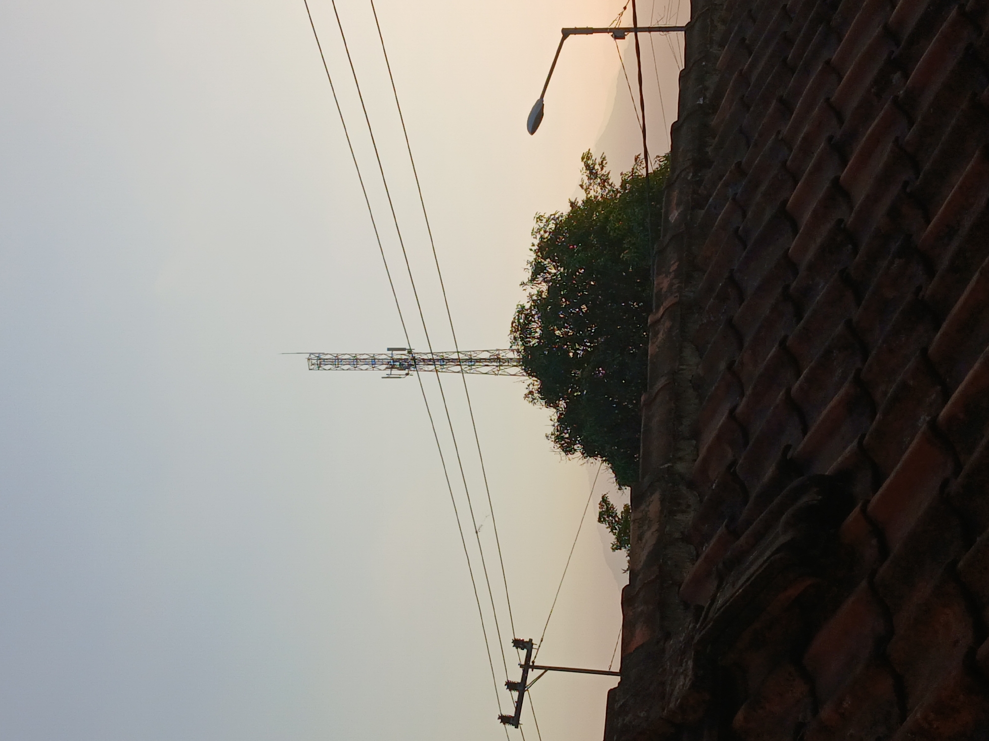 The image depicts a rooftop view in Kademangan Kulon, Indonesia, with a cell tower and power lines dominating the sky. The foreground is occupied by a weathered, tiled rooftop. In the mid-ground, a tall, lattice-style cell tower stands prominently, partially obscured by a dense, green tree. Several power lines crisscross the sky, suspended from utility poles. A street light is visible on the right side of the frame. In the background, hazy mountain silhouettes are faintly discernible against the pale sky, suggesting a rural or semi-rural setting. The lighting indicates either early morning or late afternoon. No people are present in the image.