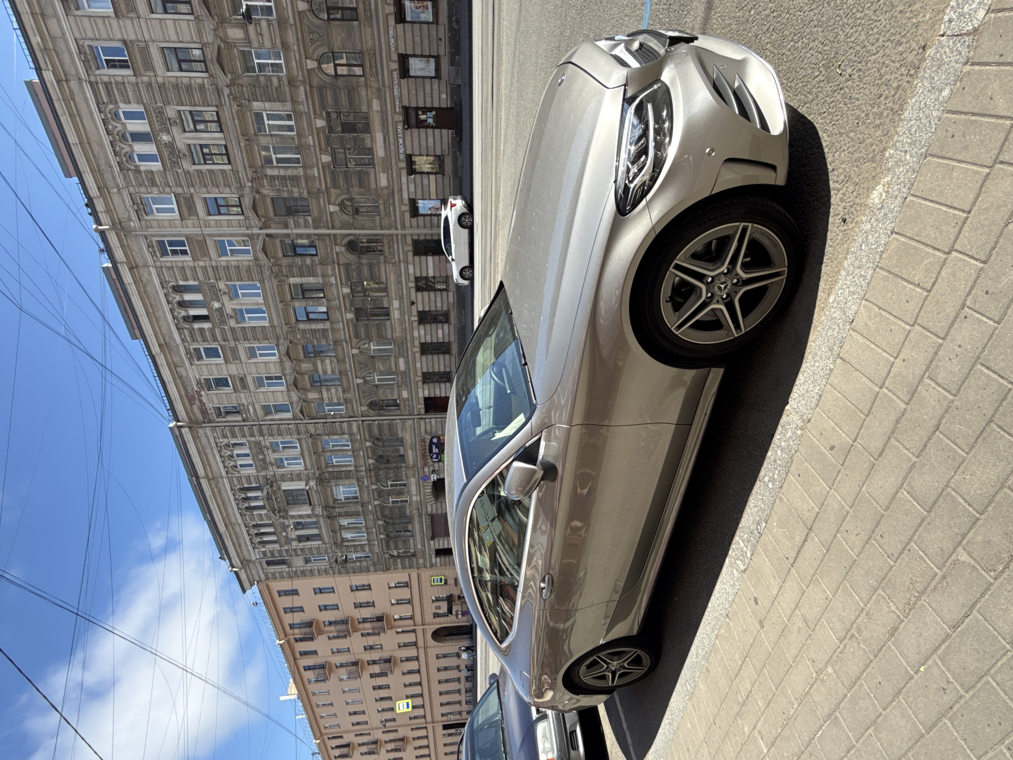 A silver Mercedes-Benz coupe is parked on a paved sidewalk along Nevsky Prospekt in Saint Petersburg, Russia. The car is in the foreground, facing right. Behind it, across the street, is a row of ornate, multi-story buildings with numerous windows and architectural detailing. A white SUV is visible driving on the street in the background. Overhead, a network of telephone and power lines crisscrosses the blue sky, which has scattered clouds. Another dark-colored car is parked to the left of the Mercedes. The scene depicts a typical urban environment in Saint Petersburg on a sunny day.