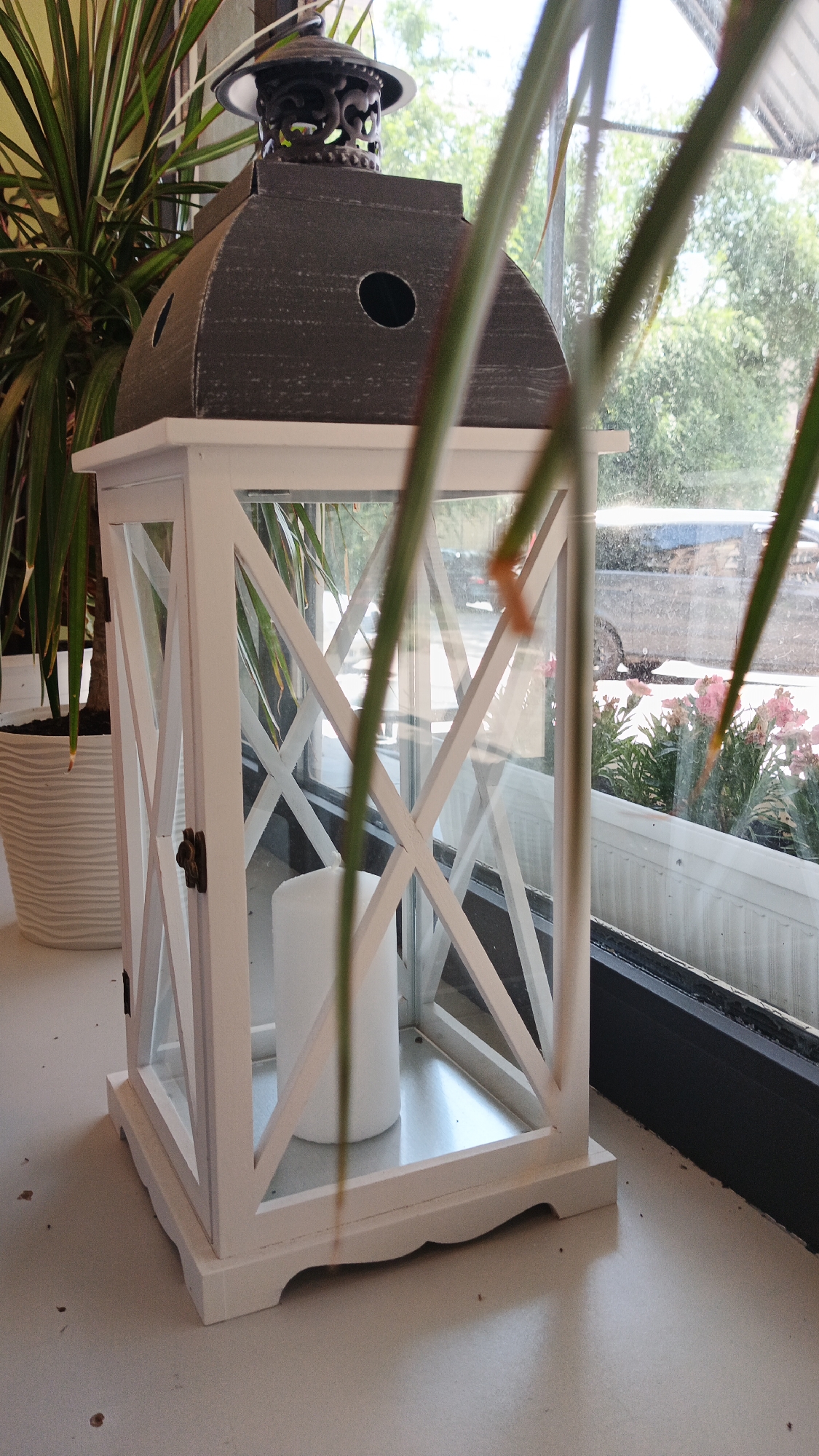 A white lantern with a dark gray top and decorative metalwork sits on a windowsill. The lantern has glass panels and white wooden lattice work. Inside the lantern, a thick white candle stands on a metal base. To the left of the lantern, a potted plant with long, green and red leaves is visible. Through the glass of the lantern and the window behind it, an outdoor scene can be seen, including trees, a car, and pink flowers in planters. The lighting suggests it is daytime. The scene is likely set indoors, by a window. There is no visible text.