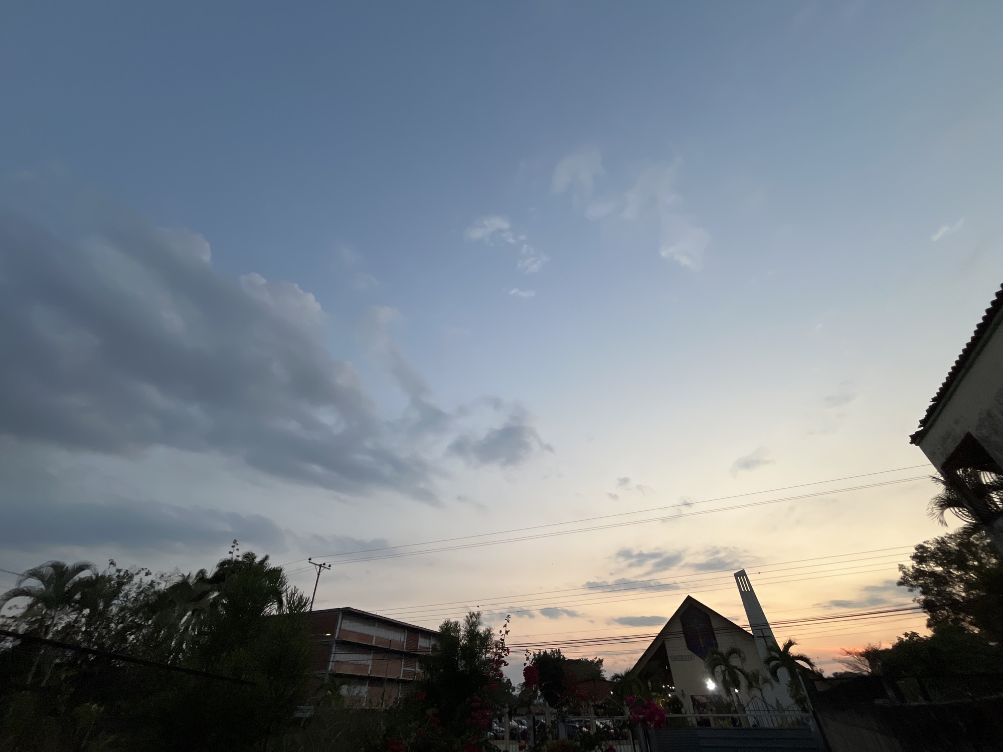 The image shows a dusk scene in Tinaquillo, Venezuela. The sky transitions from blue to orange, with scattered clouds. Silhouetted trees and rooftops are visible in the foreground. A building with a steeple, likely a church, stands out. Power lines stretch across the sky.
