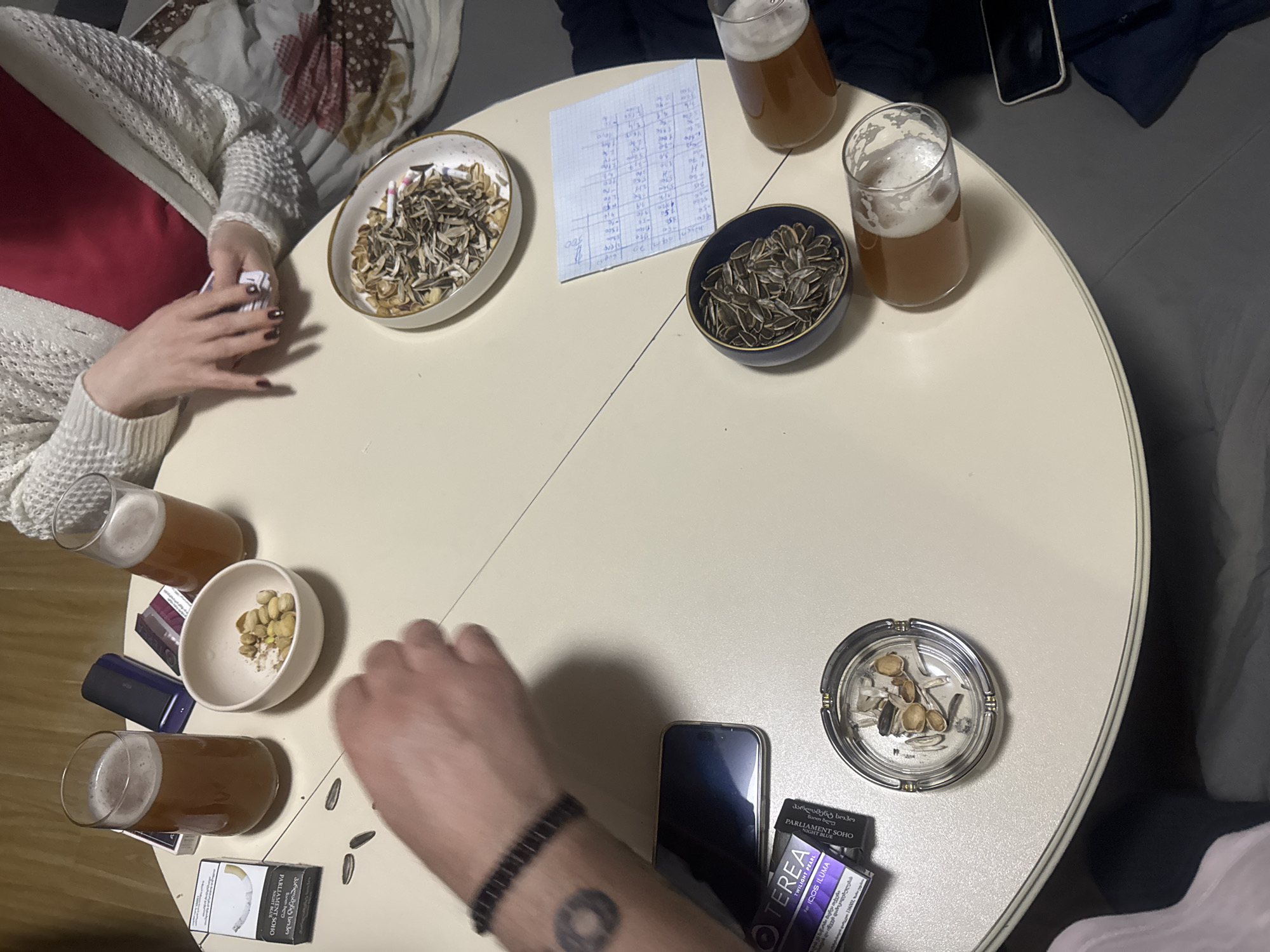 The scene depicts a casual gathering, likely in Tbilisi, Georgia, evidenced by Georgian-language cigarette packaging. The setting is a light-colored, circular table hosting four individuals, only partially visible. The table is scattered with typical social gathering items: sunflower seeds in two bowls (one light, one dark), pistachio nuts in another bowl, four glasses of beer, an ashtray containing discarded sunflower seed shells, two smartphones, cigarette packs (Parliament Soho), and a handwritten score sheet. One individual on the left wears a white knitted cardigan with a red top underneath. Another individual's arm is visible on the lower left, wearing a dark bracelet and displaying a circular tattoo. There are playing cards near that person. The general atmosphere is relaxed and informal.