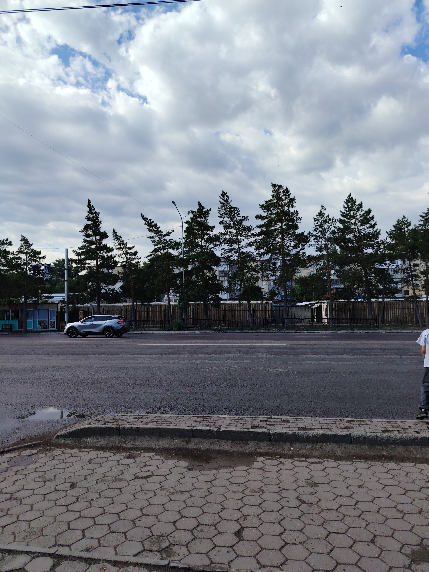 A silver SUV is driving down a city street. In the background, there are trees, buildings, and a cloudy sky. The street is paved with asphalt, and there is a sidewalk made of hexagonal tiles. A person is standing on the sidewalk, facing away from the camera. The image appears to be taken in the city of Temirtau, Kazakhstan, based on the text visible on a building in the background.