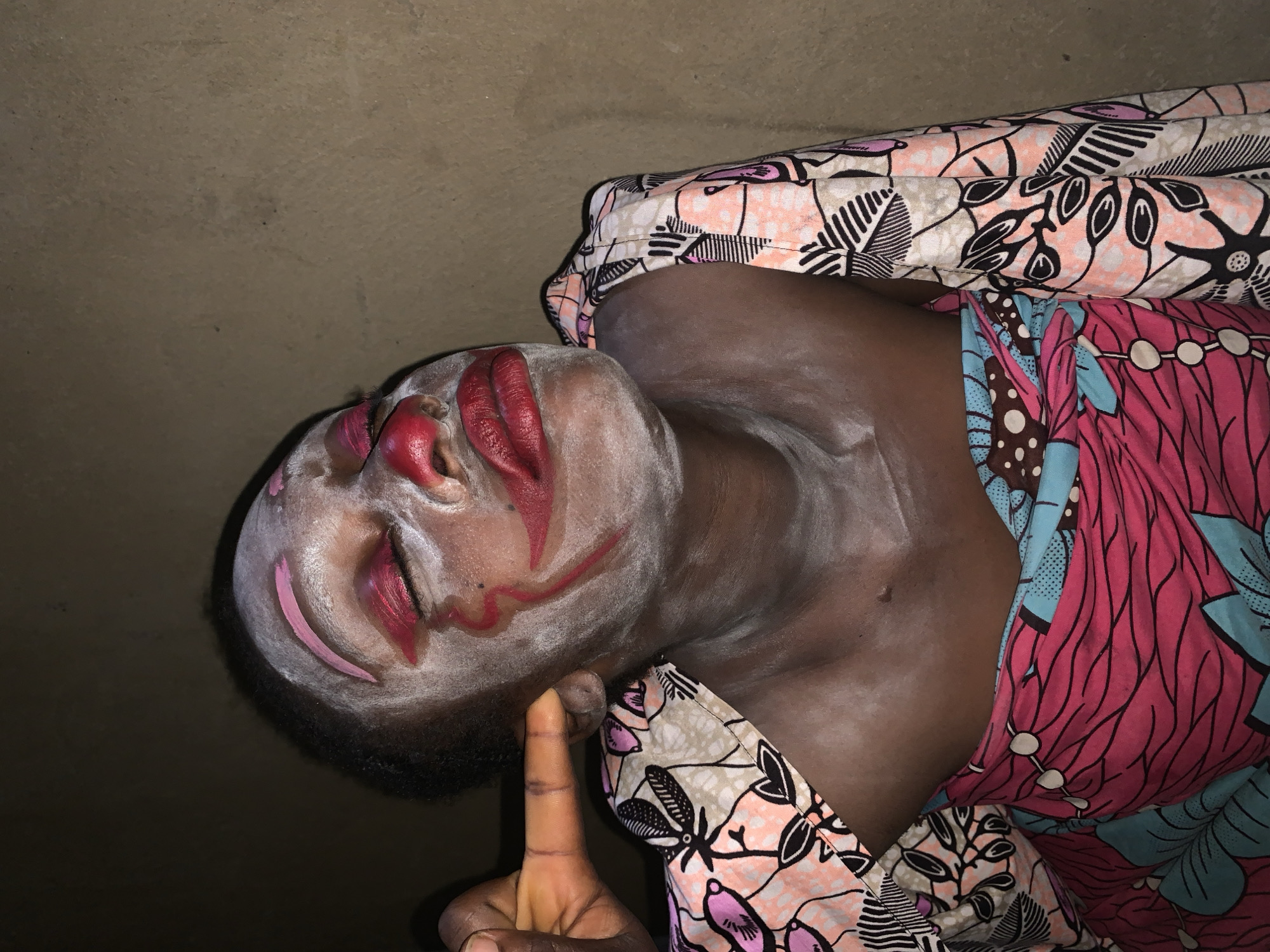 A person is seen from the chest up in a dimly lit environment. The individual has white powder applied to their face, with red and pink makeup accents on their eyes, nose, and mouth, resembling clown makeup. Pink makeup is applied as eyebrows, and red lipstick is prominently featured. A hand is touching the side of the person's face, near their ear. The person is wearing a colorful headwrap or fabric with a patterned design of pink, black, and white shapes, and a pink and blue patterned garment. The background appears to be a textured, neutral-colored wall. The setting is believed to be in Agban, Nigeria.