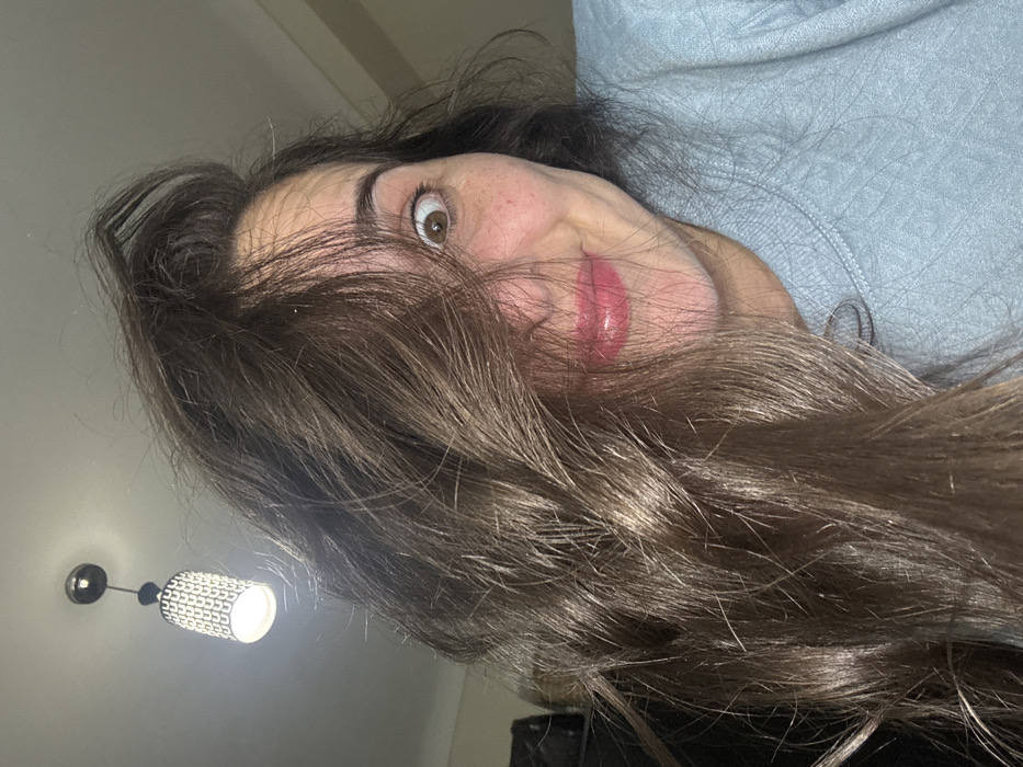 A woman lies on her back, face tilted towards the camera. She has long, wavy, light brown hair obscuring part of her face. Her right eye is wide open, with freckles visible on her cheek. Her lips are glossy. She appears to be wearing a light blue sweatshirt. A light fixture hangs from the ceiling in the lower left corner of the frame.