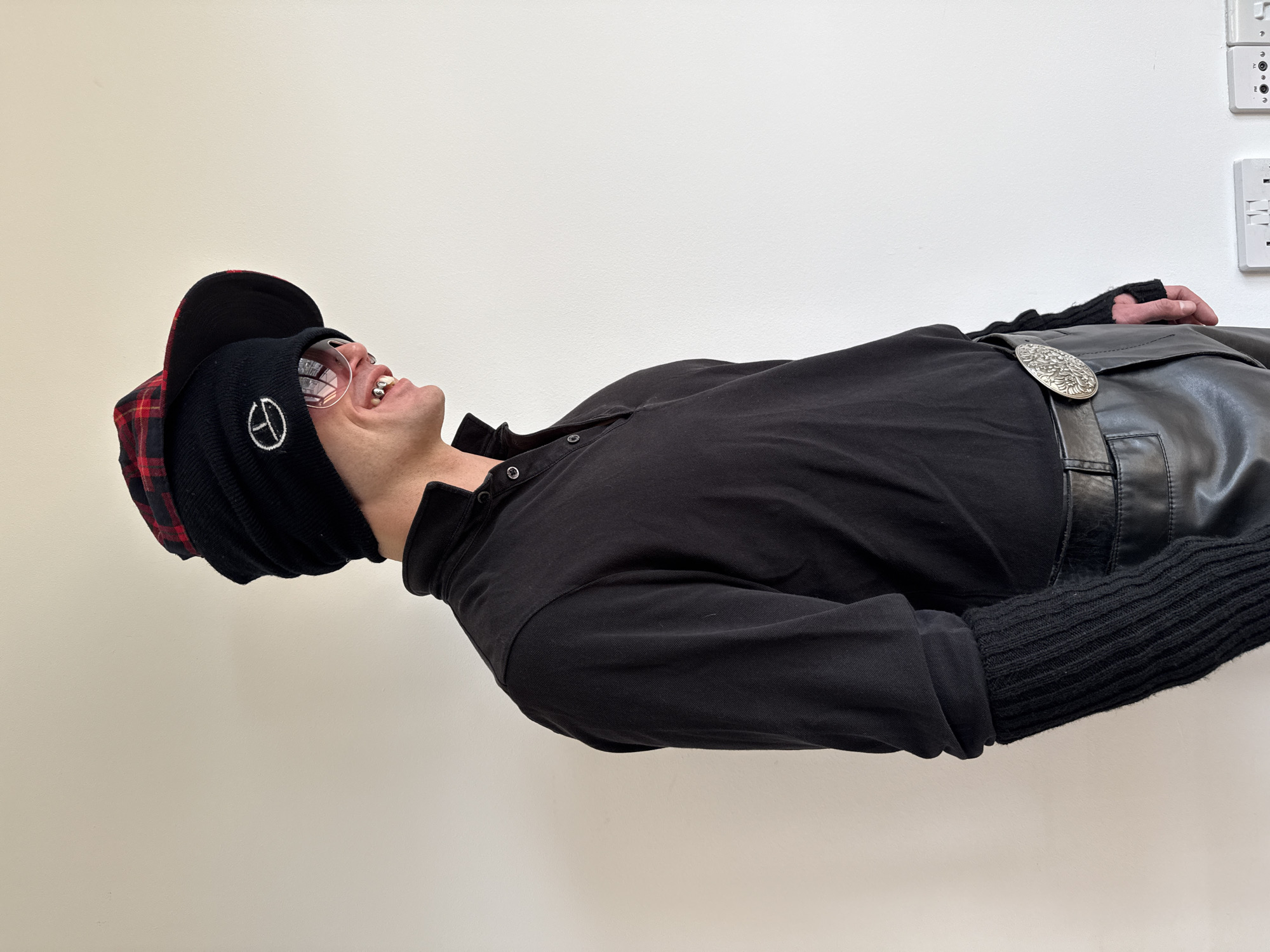 A close-up, rotated portrait features a person, likely a young adult or middle-aged male, captured against a plain, light-colored wall. The individual is looking upwards with an open-mouthed smile, revealing distinctive metallic caps or grills on several upper teeth.
The person is dressed in a black long-sleeved top. On their head, they wear a black knit beanie adorned with a white peace sign emblem, with a red and black plaid baseball cap visible either beneath or over it. Around the waist, a black leather-look garment, possibly a kilt or overskirt, is fastened by a prominent, ornate silver buckle. The left arm, from the viewer's perspective, is visible, covered by a black fingerless knit glove or arm warmer.
The background is a simple, light-colored wall, with white UK-style electrical sockets partially visible in the upper right corner. This detail confirms an indoor setting and aligns with the provided location context of London, United Kingdom. The overall expression of the person conveys happiness, amusement, or a playful mood.