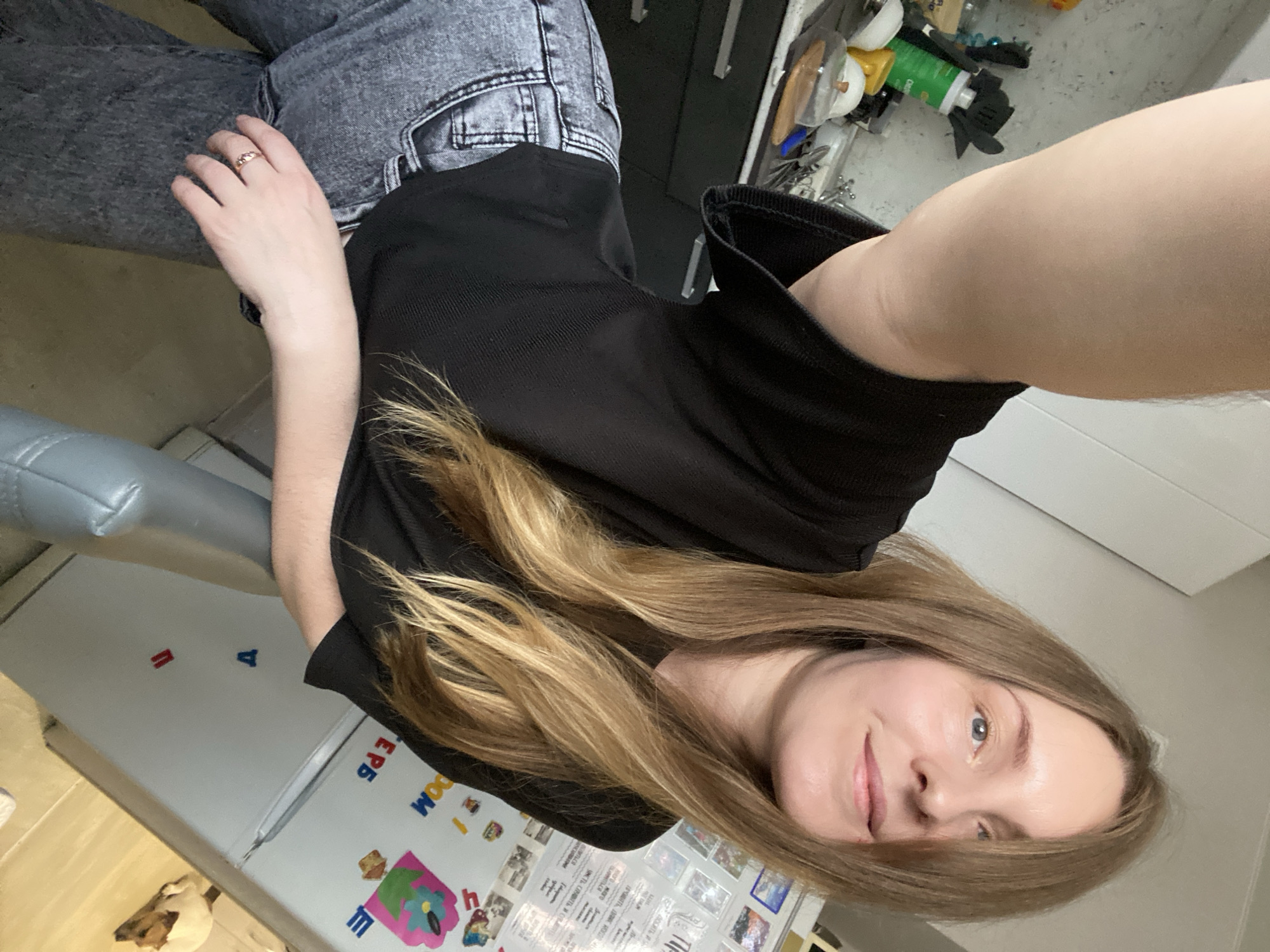 A woman with long blonde hair and blue eyes is shown in a selfie-style image. She is wearing grey jeans and a black top. A ring is visible on her left hand. In the background, there is a refrigerator covered with magnets and pictures. The image was taken at an angle, so the specific location in the city of Noviy, Russia is not discernible. Other background objects include a grey chair or ottoman and a storage cabinet with items on top of it.