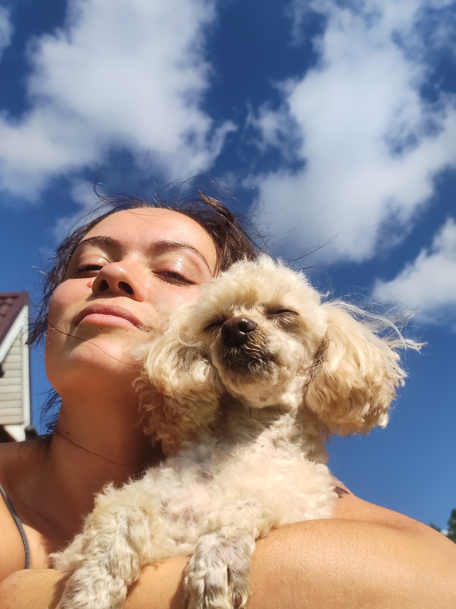The image is a low-angle, close-up shot of a young woman holding a light-colored, fluffy dog against a bright blue sky with white clouds. The woman's eyes are closed, and she appears to be relaxed, with sunlight illuminating her face. The dog, also with closed eyes, is nestled against her chest. The background shows a hint of a house with a brown roof to the left and some green foliage on the right. The scene is outdoors and appears to be a sunny day. The location is identified as the city of Fokino, Russia.