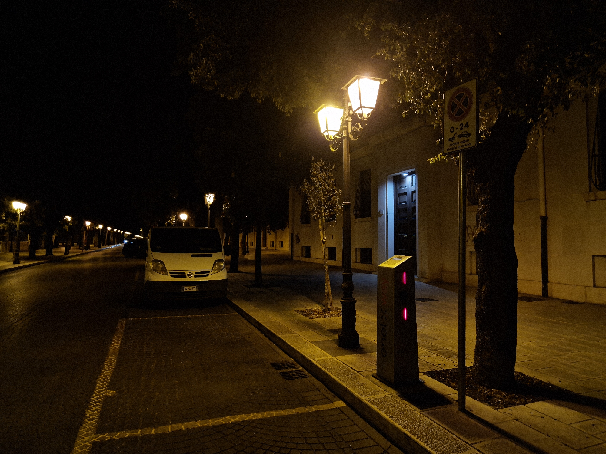 A nighttime, low-light street scene in Taranto, Italy, captured from a street-level perspective. The image is dominated by the warm glow of streetlights illuminating a tree-lined street and sidewalk. A white van is parked on the left side of the street, facing away from the camera. To the right of the van, a modern, illuminated charging station for electric vehicles is visible, bearing the "enelx" logo and showing two red indicator lights. Further to the right, a historic-style lamppost with multiple lights stands on the sidewalk. A traffic sign indicating parking restrictions (no stopping, 0-24 hours, for electric vehicle charging) is mounted on a pole near a large tree. The street and sidewalk are paved with cobblestones and tiles. Buildings with illuminated doorways are visible in the background. The overall atmosphere is quiet and serene, characteristic of a city street at night.