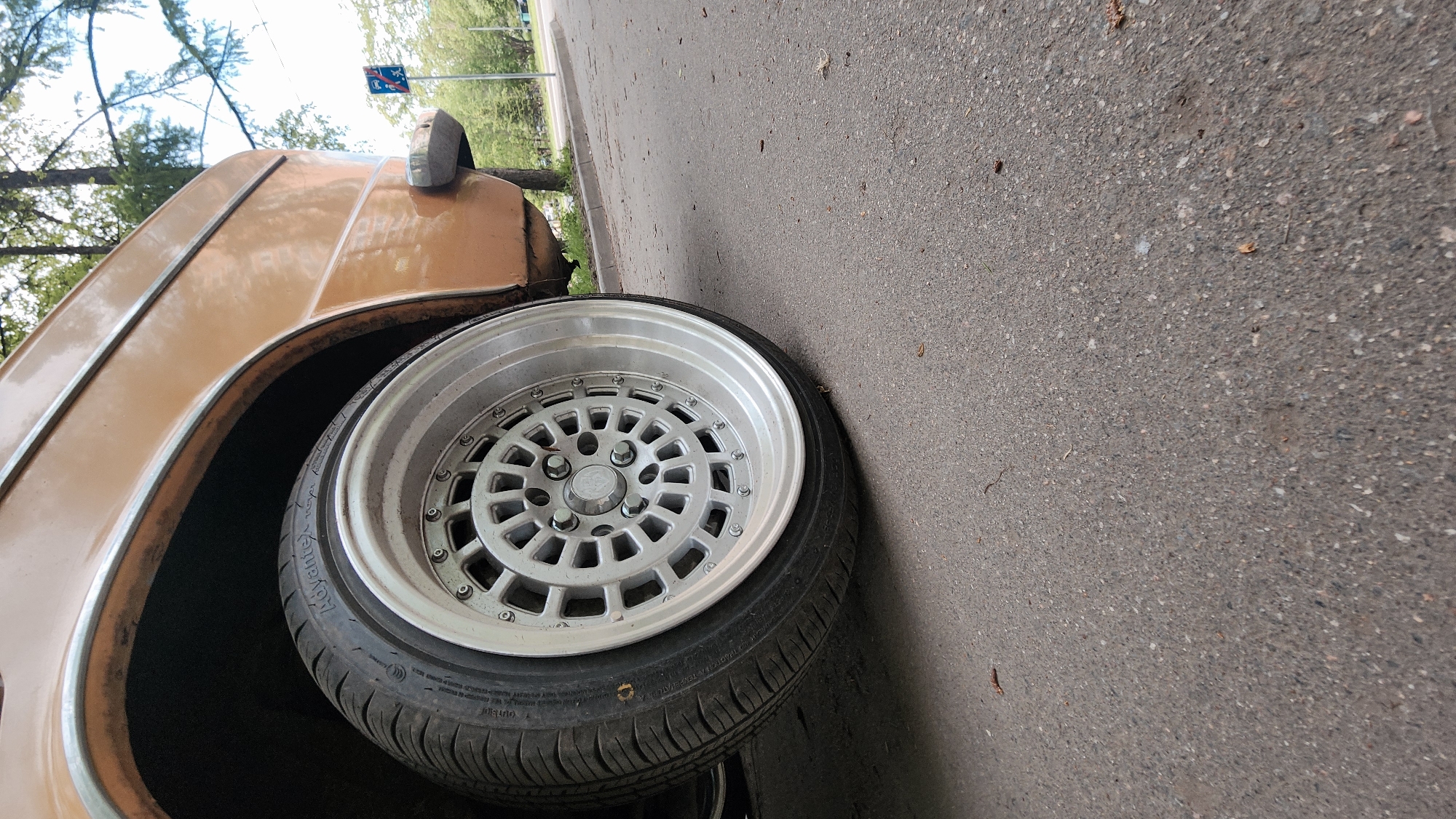The image shows a close-up view of a car wheel, specifically the front passenger side, resting on an asphalt road. The car appears to be an older model, possibly from the 1970s or 1980s, with a faded brown or gold paint finish. The wheel has a custom silver rim with a complex, multi-spoke design. The tire is black, with the word "Advanti" visible on the sidewall. A small portion of the car's fender is visible above the wheel, showing some rust and wear. In the background, blurred trees and a street sign are visible, suggesting an urban or suburban environment. The image appears to be taken in Moscow, Russia, because the sign visible in the background has the flag of the Moscow region. The asphalt road is typical for urban road construction in Moscow. The overall condition of the car and the setting implies the image was taken in a less affluent area. The angle of the photo suggests it was taken at ground level, possibly by someone crouching down.