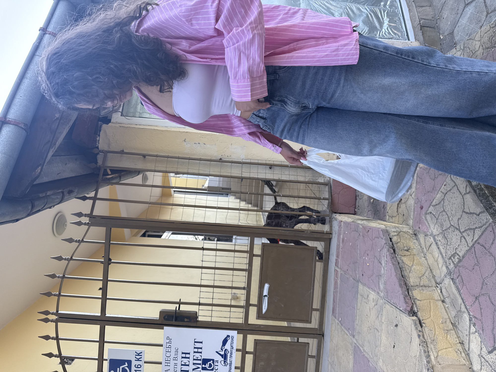 A young woman with curly dark hair, wearing a white tube top, a pink and white striped open button-down shirt, and blue denim jeans, stands on a cracked tiled walkway outside a building. She holds a white plastic bag in her right hand. In the background, a metal gate is slightly ajar, revealing a dark-colored speckled dog peeking through it. Above the gate, a sign with text and symbols is visible. Part of the building's facade, a slanted roof, and a metal railing are also in the frame. The environment appears to be a street in Sveti Vlas, Bulgaria, characterized by the tiled pavement and building architecture.