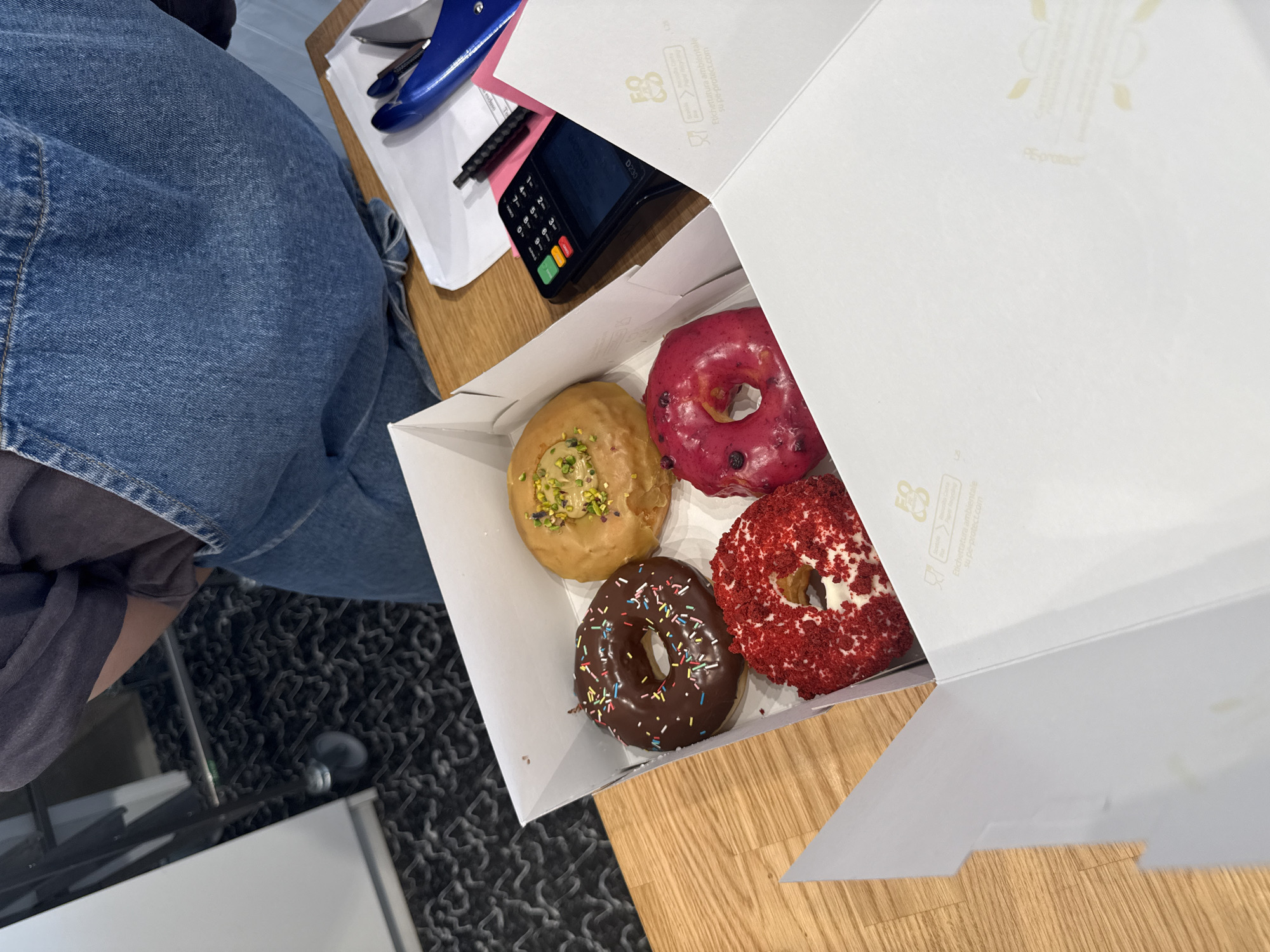 An overhead shot captures an open white box containing four assorted donuts, placed on a light wooden table. The donuts feature various toppings: one light brown with green and yellow sprinkles (possibly pistachio), one bright pink/red with dark berry pieces, one dark chocolate with colorful rainbow sprinkles, and one red with red crumbs (resembling red velvet).
To the left of the donut box, part of a person is visible, wearing blue denim jeans and a dark grey or black top. On the table near the person, a blue credit card terminal, labeled "D200," is visible with its keypad and colored function buttons. Next to the terminal are a black pen and some white papers, with a blue office tool (possibly a stapler or cutter) lying on top of them.
The donut box itself has text printed on its inside lid: a stylized logo, followed by "Emballage alimentaire / Paper food packaging / PE-protect" and a web address "peprotect.com." The table's wooden surface takes up the right side of the frame, while a dark, patterned carpet or rug is visible on the bottom left. The scene suggests an indoor setting, possibly a cafe or bakery counter, in Milan, Italy.