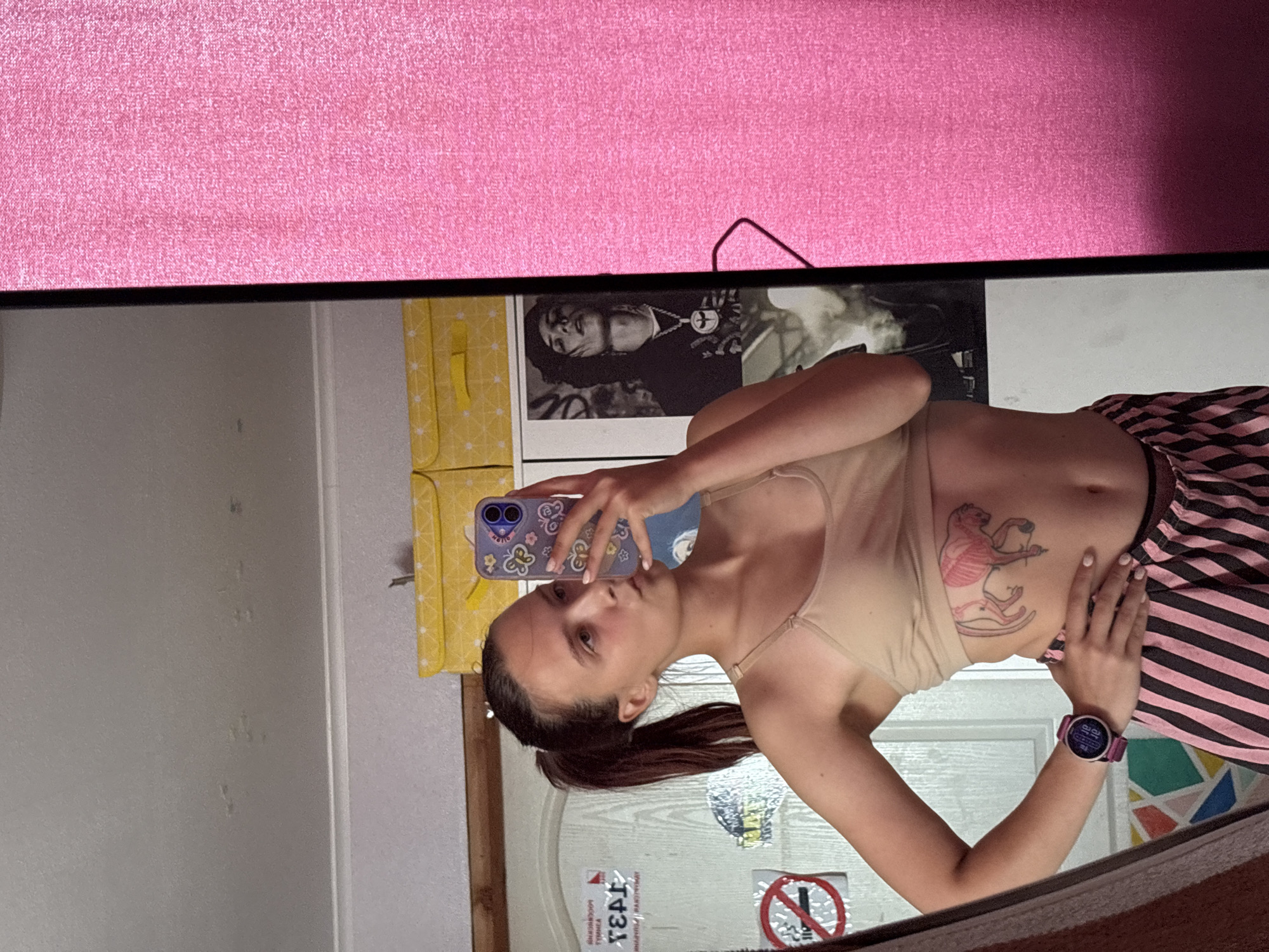 The image is a medium shot of a young woman taking a mirror selfie in what appears to be a bedroom. The woman has brown hair pulled into a ponytail and is wearing a beige tank top and pink and black striped pajama pants. She has a tattoo on her left side. She is holding a purple phone with butterflies on the case. In the background, there is a door with stickers, some yellow storage boxes on a shelf, and a poster of a man with dark hair. A pink curtain is visible on the right side of the frame. The overall scene suggests an indoor, personal space. The location is unknown.
