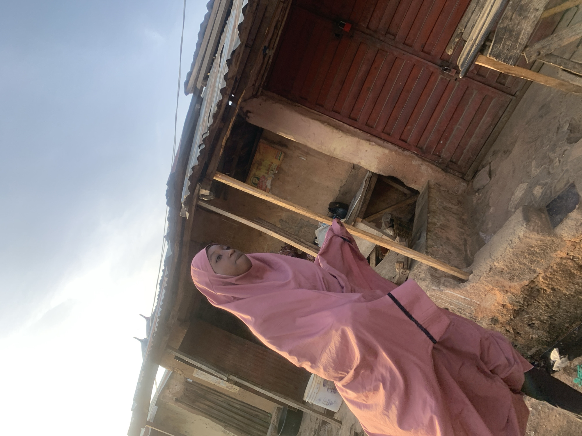 A medium shot captures a person wearing a pink hijab and long dress, standing in what appears to be a modest, possibly older part of the city of Jos, Nigeria. The backdrop reveals weathered buildings, some with corrugated iron roofing and wooden supports. One building has a red wooden door secured with a padlock. The overall setting is outdoors, under a bright sky.