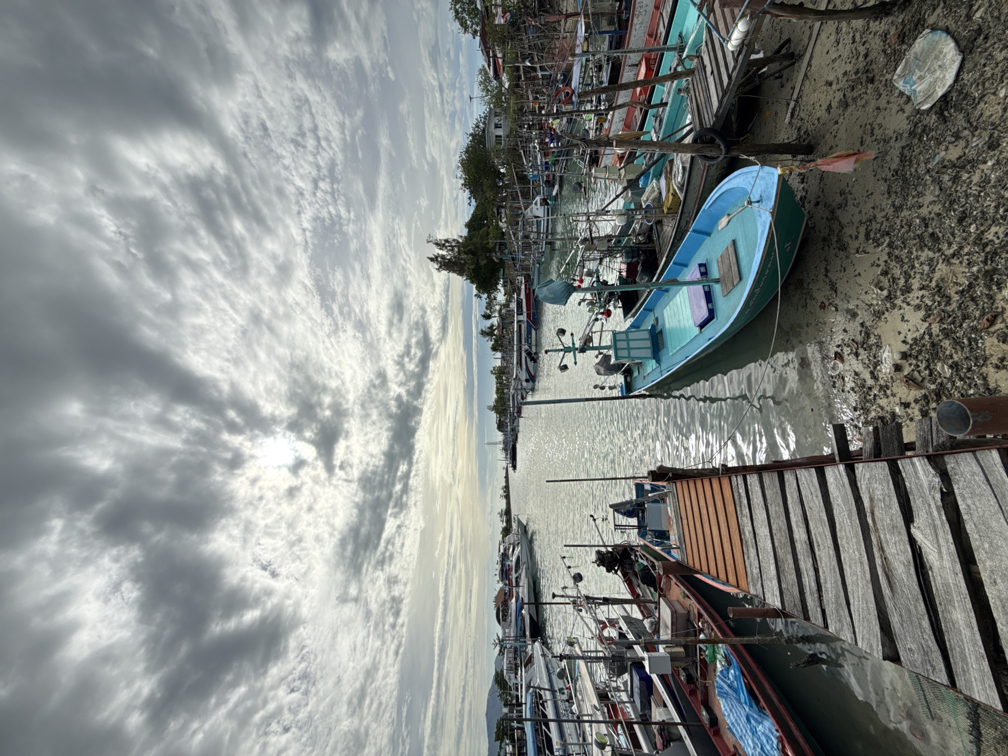 The image depicts a harbor on the island of Ko Samui in Thailand, under a cloudy sky. Numerous fishing boats, a mix of small and medium-sized vessels, are docked along a sandy shore and a wooden pier. The water appears calm, reflecting the overcast sky. Some boats are painted in various colors, including blue and green, while others are more weathered. Fishing equipment and netting are visible on some of the boats. The shore is covered with sand, mud, and debris, suggesting a working harbor. In the background, trees and buildings are visible, indicating the presence of a settlement. The overall scene is that of a typical fishing village harbor.