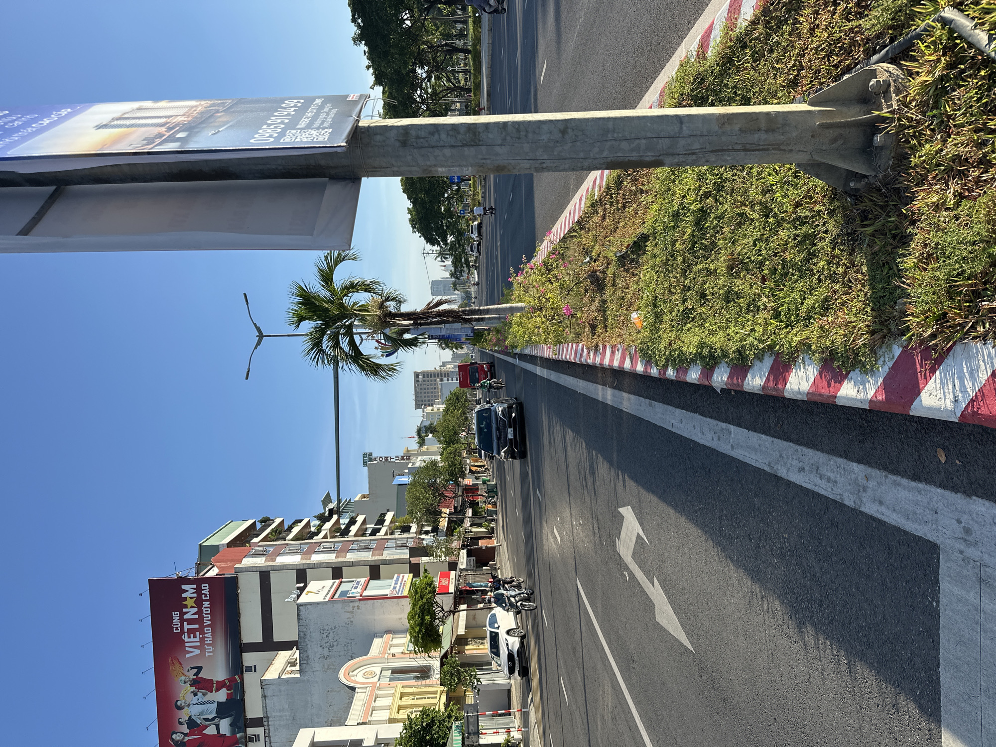 The image captures a street scene in Da Nang, Vietnam, likely in the early morning or late afternoon due to the long shadows. The focus is on a multi-lane road with traffic. On the left side of the road, there are buildings, including one with a large red billboard featuring people and text in Vietnamese. Further down the street, white and black cars are visible, along with motorcycles. A white car is parked on the side of the road, and a person on a motorcycle is in motion.
The right side of the road features a median strip with palm trees and greenery. A large utility pole stands prominently in the foreground, supporting a sign that appears to be an advertisement. The road itself has clear lane markings, including a left-turn arrow painted on the asphalt. The sky is clear and blue. The overall environment suggests a well-developed urban area with ongoing traffic and commercial activity.