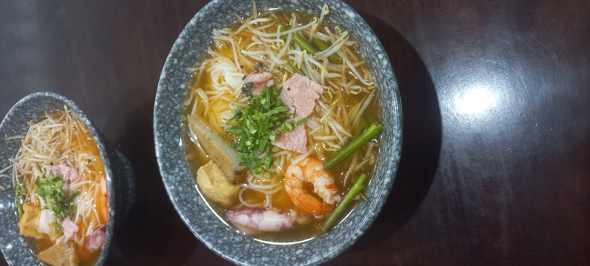 Two bowls of Vietnamese noodle soup, likely pho, are presented on a dark wooden table. The bowls are grey with a speckled pattern. The soup contains noodles, bean sprouts, sliced beef, shrimp, calamari, and pieces of tofu. Chopped green onions garnish the top of the soup in the foreground bowl. The lighting suggests an indoor setting, possibly a restaurant, and the overall impression is that of a prepared meal ready to be served or consumed. The scene does not provide any specific indicators of its location within Da Nang, Vietnam, beyond the implied context of Vietnamese cuisine.