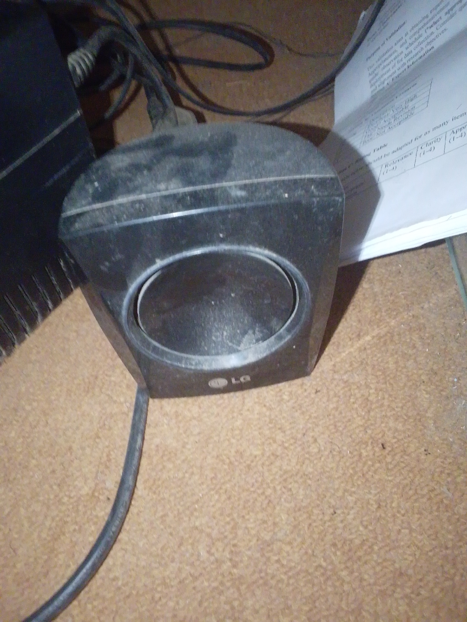 The image shows a close-up, eye-level shot of a small, black LG speaker. The speaker is dusty, with visible grime on its surface, particularly around the speaker cone and the LG logo. The speaker is positioned on a brown, carpet-like surface. To the left of the speaker, a portion of a larger black electronic device is visible, along with several black wires and cables. In the background, a white sheet of paper with printed text is partially visible, suggesting a workspace or study area. The lighting appears to be artificial and somewhat dim, casting soft shadows. The focus is sharp on the speaker, with the background slightly blurred. No people or specific events are depicted. The text on the paper includes phrases like "Purpose of Validation," "appropriateness," and a table with headings like "Relevance" and "Clarity," indicating a context related to data collection or research. The location context of Jalingo, Nigeria, does not provide any specific visual cues in the image itself.