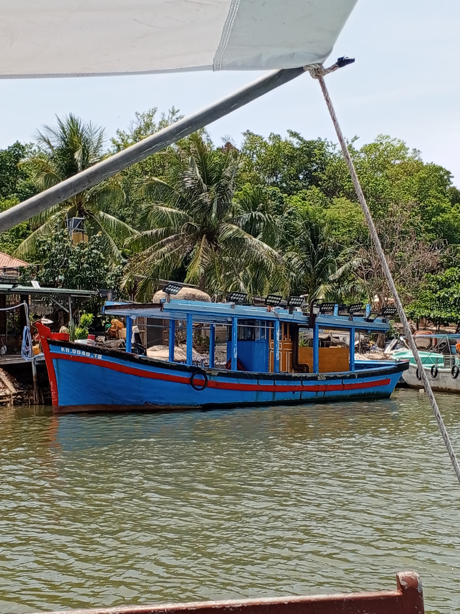 A blue and red fishing boat is docked on the right, in the city of Bac Nha Trang Ward, Vietnam. The boat is equipped with a roofed seating area and multiple floodlights on the roof. Palm trees and lush green foliage line the background, suggesting a tropical environment. The water in the foreground is calm and appears murky green. Another white boat is partially visible to the right of the main fishing boat.