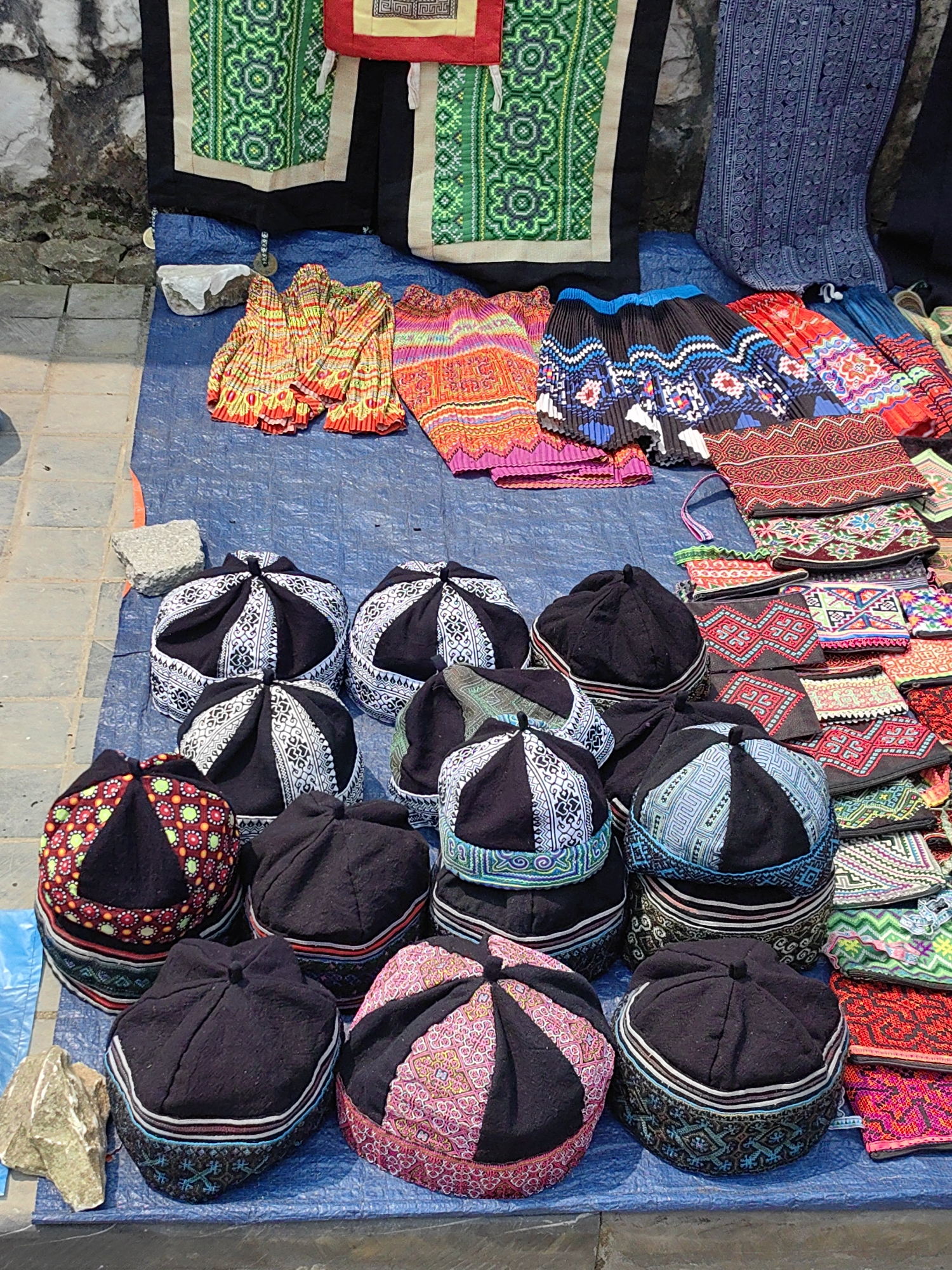 The scene captures a market stall in Sa Pa, Vietnam. Textile goods are displayed outdoors on a blue tarp spread over a paved surface next to a stone wall. In the foreground and midground, a collection of approximately 16 dome-shaped hats are arranged on the tarp. These hats feature black fabric bodies with embroidered panels on their crowns and sides, exhibiting patterns in colors such as white, black, red, pink, green, and blue. Behind these hats, various pleated textile garments, likely skirts, are folded on the tarp, showcasing multi-colored patterns including bright oranges, pinks, blues, and blacks. To the right, a stack of smaller, rectangular embroidered pouches or bags, displaying geometric patterns in reds, greens, blues, and other colors, is present. Hung vertically against the gray stone wall in the background are larger textile items with prominent green and black geometric embroidery, and a piece of deep blue patterned fabric. A rough-textured rock rests on the blue tarp on the left side of the display. No individuals are visible in the image.