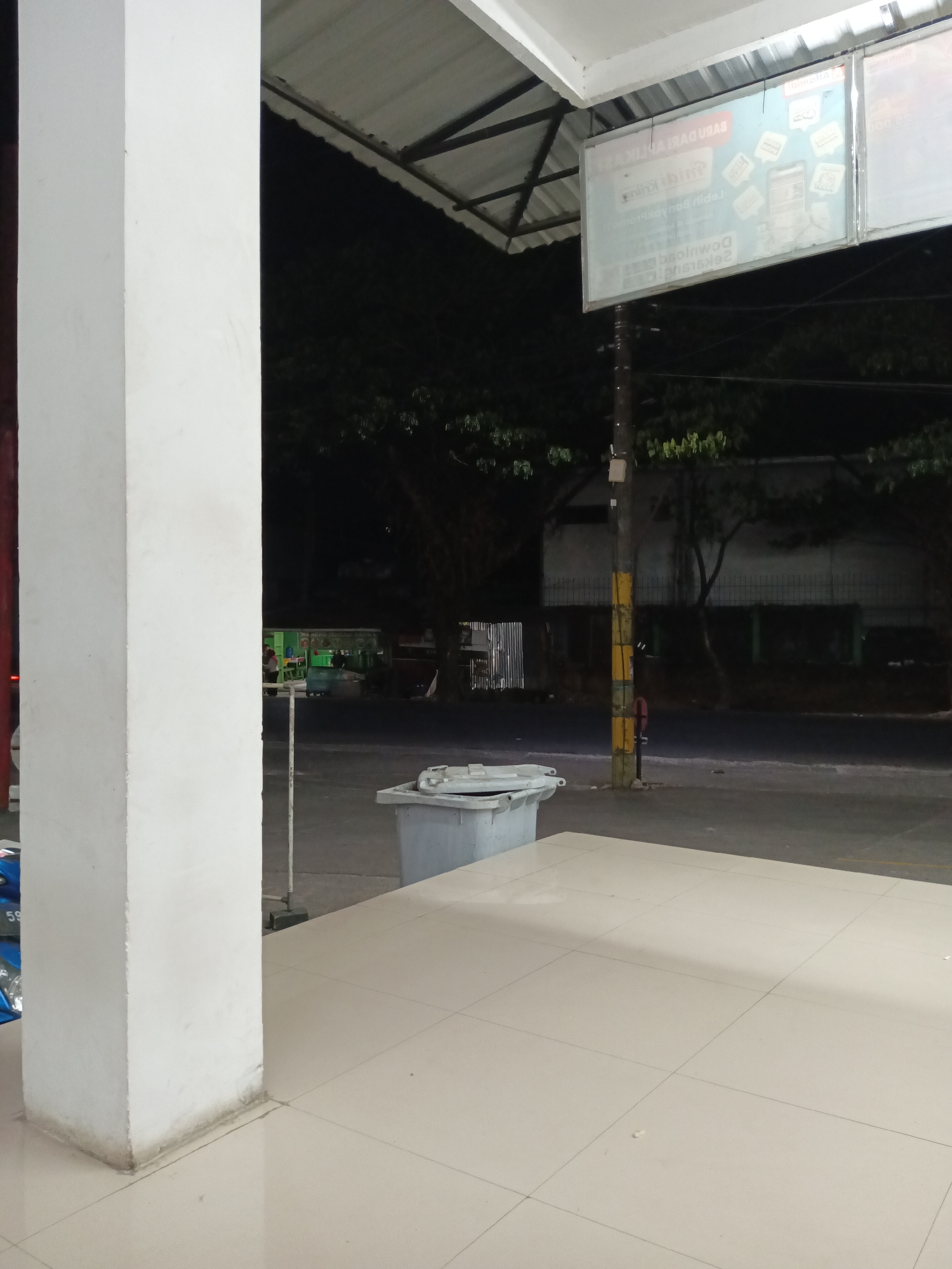 This image depicts an outdoor scene at night, likely in Makassar, Indonesia, given the provided context. In the foreground, a large white concrete pillar dominates the left side of the frame. The ground is tiled with large, light-colored square tiles. To the right of the pillar, a light grey dumpster is visible. Further back, a street is visible with a pole marked with yellow and black stripes. Beyond the street, there are some trees and a building with what appears to be a green facade. A sign is suspended above the scene, illuminated and displaying text and graphics, though the details are not clearly legible. The overall impression is that of a public area or street scene at night.
