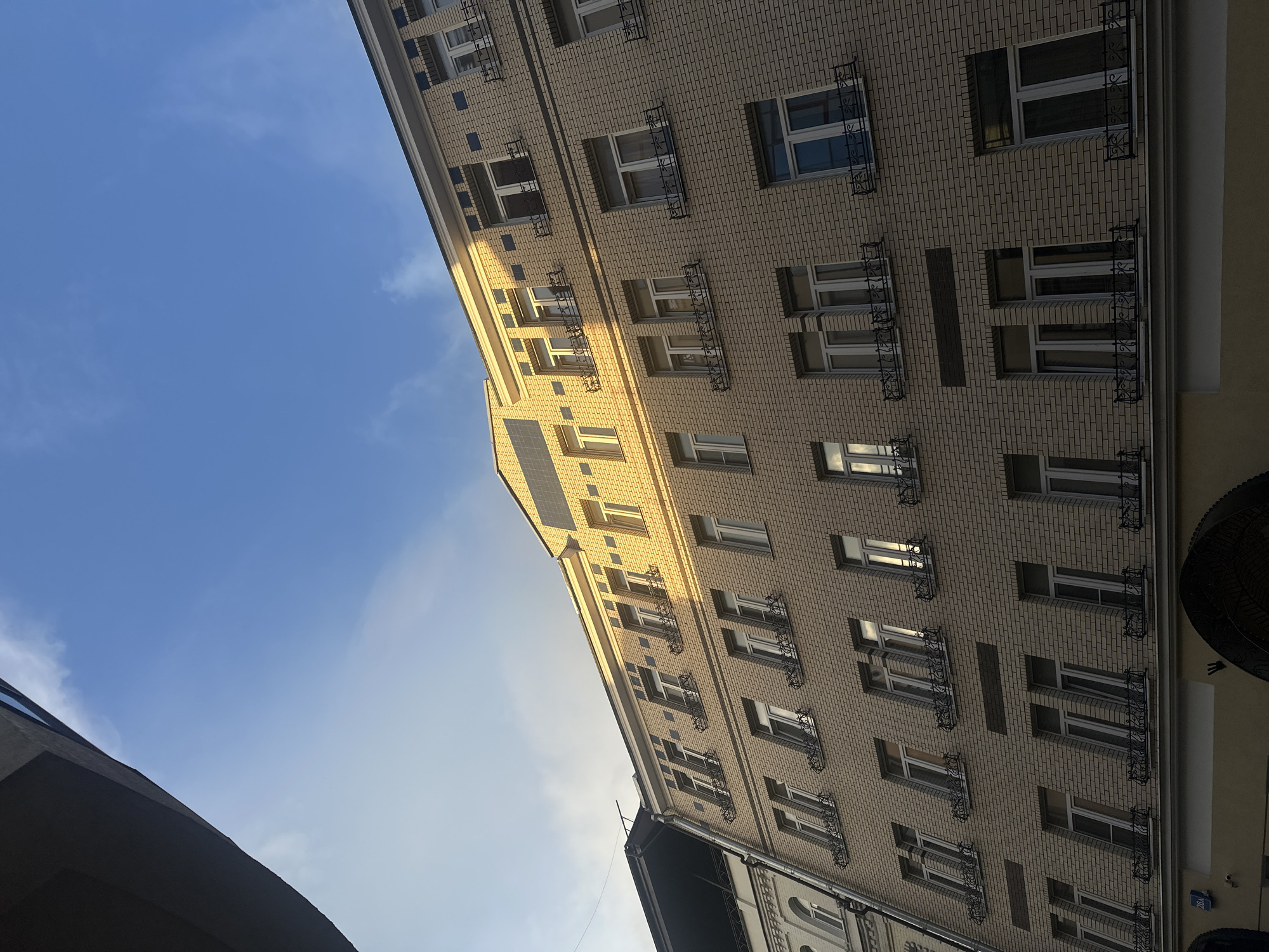 The photograph captures the façade of a light-yellow brick apartment building under a clear blue sky with scattered wispy clouds. The building features multiple stories with numerous windows, many adorned with decorative black wrought-iron railings and some with small window boxes. The architecture suggests a European style, possibly from the early to mid-20th century, with decorative elements above some windows and along the roofline. Sunlight illuminates the upper sections of the building, casting shadows and highlighting the texture of the bricks. A dark, angular structure, possibly another building or overhang, is visible in the upper left corner, creating a stark contrast with the sky and the lit portion of the apartment building. The perspective is from a low angle, looking upwards towards the building and the sky. The scene is devoid of people or any overt human activity. A blue street sign with the number "26A" is partially visible at the bottom left of the building. Based on the architectural style and the street sign, this building is located on Patriarch's Ponds in Moscow, Russia.