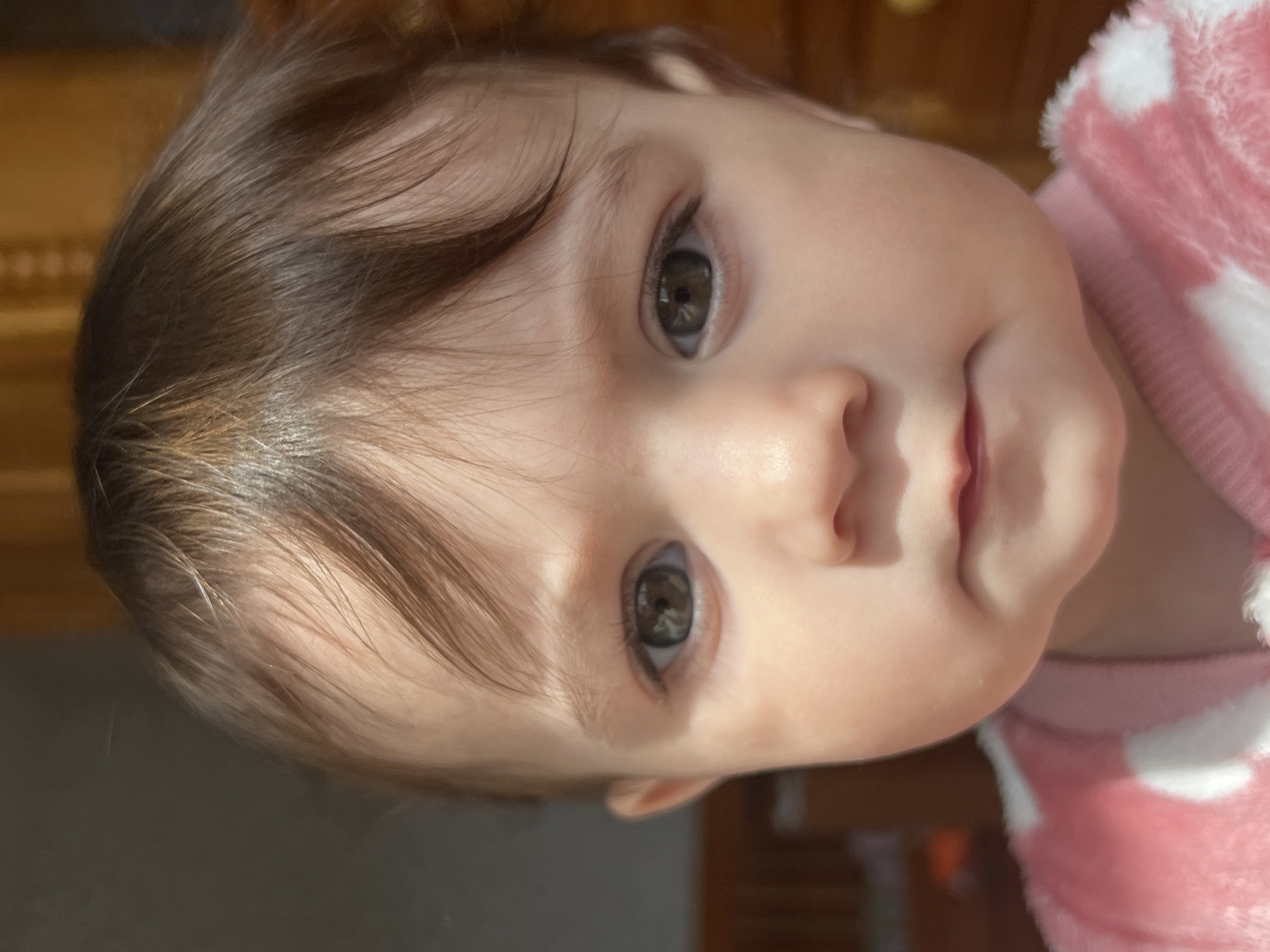 A close-up, slightly tilted portrait of a baby with fair skin, brown hair, and hazel eyes is captured. The baby is wearing a pink and white polka-dot outfit. Natural light is illuminating the scene, creating highlights on the baby's face. The background is blurred, but suggests an indoor setting with wooden furniture or paneling visible.