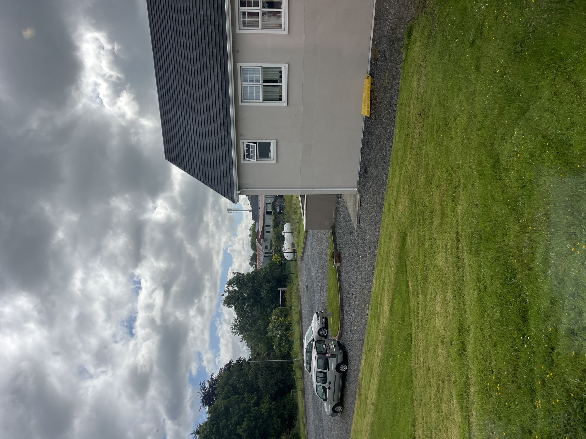The image depicts a scene in Tyrrellspass, Ireland, on a cloudy day. In the foreground, a well-maintained green lawn slopes upwards. Beyond the lawn, a gravel parking area is visible, containing two parked cars: a silver minivan and a silver sedan. To the right, the side of a light-colored building with a dark, tiled roof is prominent, featuring white-framed windows. A bright yellow object, possibly a traffic barrier or a construction item, lies on the gravel near the building. In the background, a line of trees borders the parking area, and further back, several buildings are visible, including a large white cylindrical tank that appears to be a propane tank, and some houses with red roofs. A cellular tower stands tall in the distance. The sky is filled with a mix of white and gray clouds.