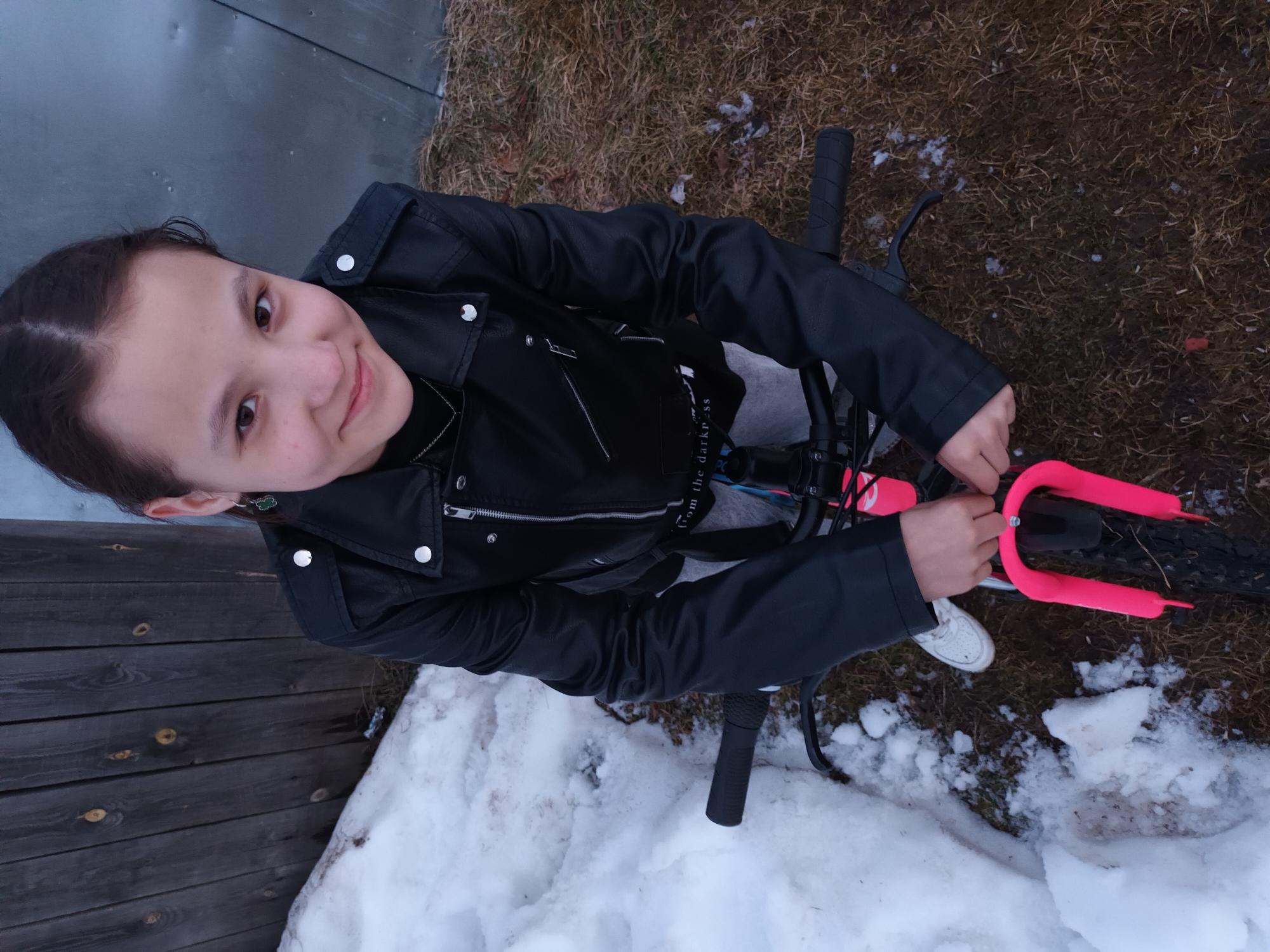 An outdoor scene captures a young female, estimated to be a pre-teen or early teen, positioned behind the handlebars of a bicycle. She has medium-brown hair pulled back and wears a black faux-leather motorcycle jacket with silver snap buttons and zippers, a dark top with a faint "from the darkn_ss" inscription, light-colored pants, and white sneakers. A green earring is visible in her left ear. Her hands are placed on the black handlebars and the bright neon pink front fork of the bicycle. She maintains direct eye contact with the viewer and exhibits a slight smile. The ground on the left side of the frame is covered in melting snow and ice, while the right side displays patches of dry, matted brown grass and dirt. In the background, a weathered wooden plank fence is visible to the left, and a light grey, possibly corrugated metal sheet with visible dents, forms the background to the right. The image does not contain visual identifiers specific to the city of Ольховка, Russia; however, the environmental conditions suggest a temperate region experiencing a thaw.