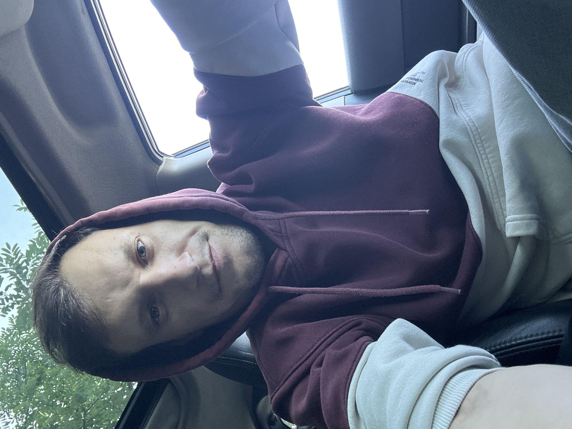 A man wearing a burgundy and cream hooded sweatshirt sits in the driver's seat of a car, looking directly at the camera. He has short brown hair and a light beard. The car has a sunroof open, with green foliage visible through it, suggesting an outdoor setting. The man is positioned in front of the dashboard and car interior, with the window and sky visible to his right. The overall impression is a selfie taken from inside the car. The location is Makiv, Ukraine.