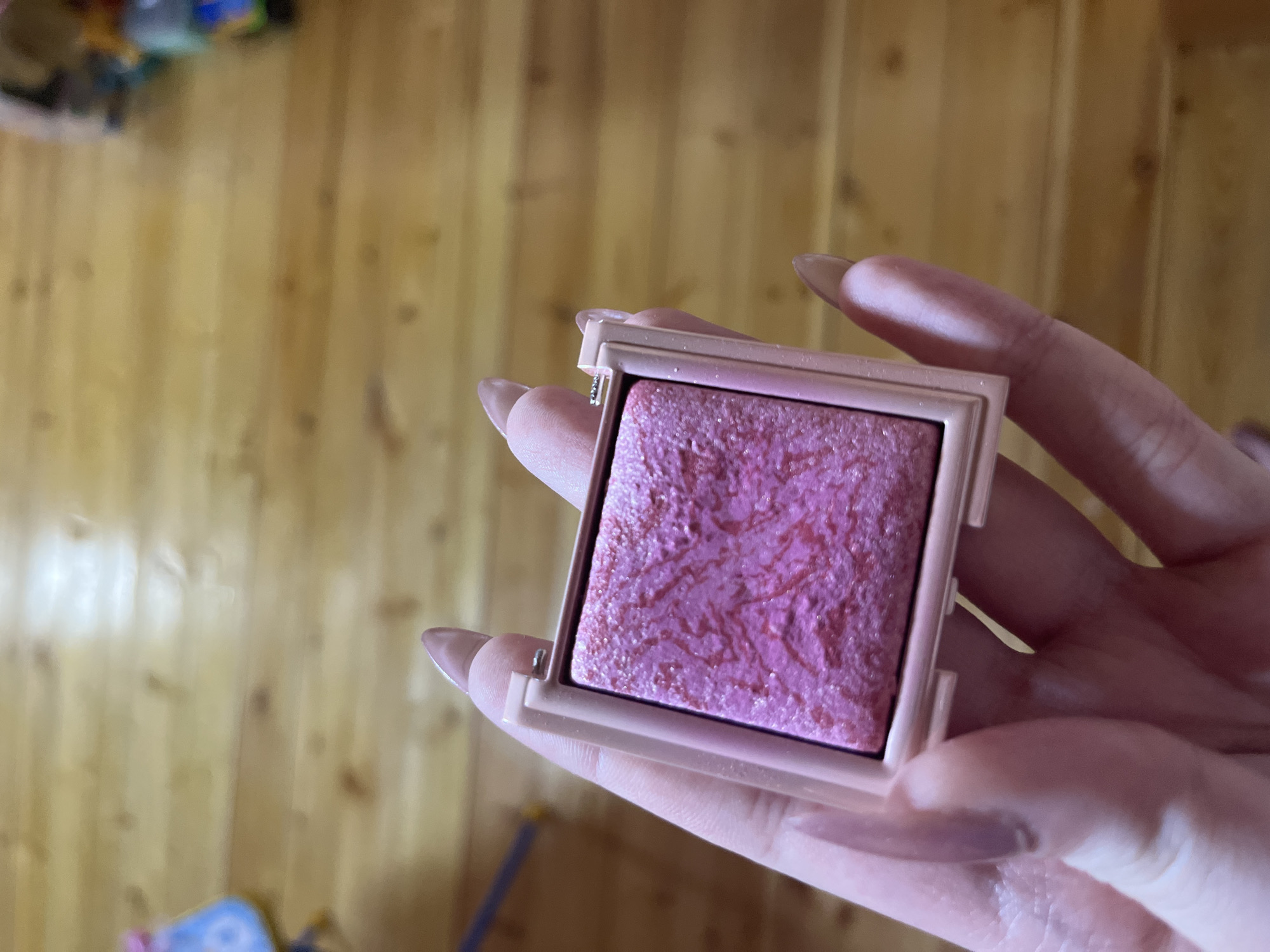 A hand with long, light-colored acrylic nails holds a square compact of shimmery pink blush. The blush has a marbled pink pattern. The compact has a light pink casing. The background is a wooden floor. The image is cropped to focus on the hand and blush. The location in Ashkadar, Russia, is not discernible from the image.