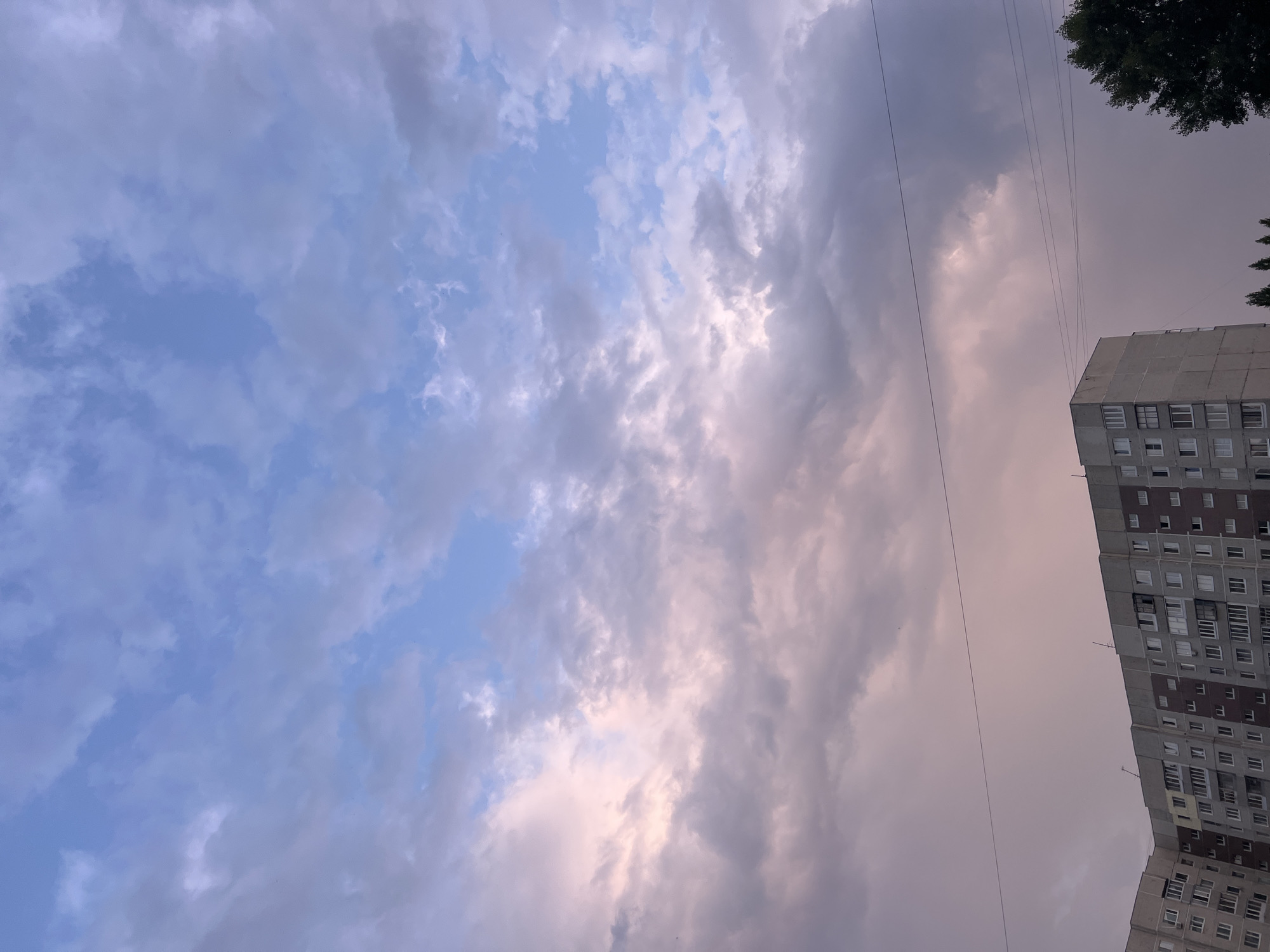 The image depicts the sky above an apartment building in Ulyanovsk, Russia. The sky is filled with a mix of light blue and fluffy clouds, with some clouds tinged with pink and purple hues, suggesting either sunrise or sunset. A large, multi-story apartment building with numerous windows dominates the lower portion of the frame. Utility wires are visible stretching horizontally across the sky. A dark green tree is partially visible on the right side of the image. The overall scene captures an urban environment with a dramatic sky.