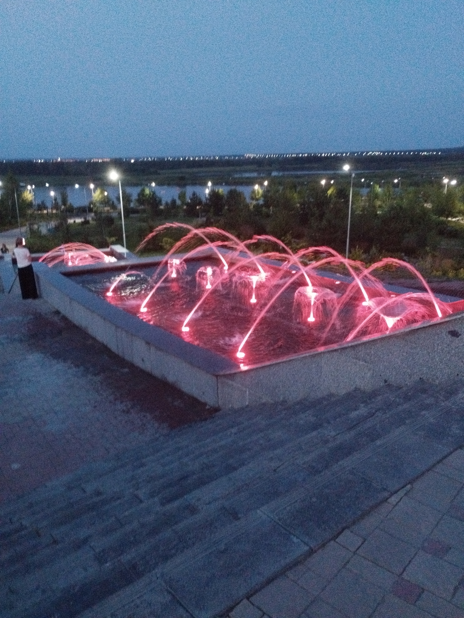 The image shows a public fountain at dusk or night, illuminated by bright red lights. The fountain is in a rectangular pool with a concrete surround, and water jets arc upwards and outwards, creating a dynamic display. The water itself appears pinkish due to the artificial lighting.
In the foreground, a set of stone stairs leads down from the viewer's perspective. On the left side of the fountain, two people are visible. One, wearing a light-colored top and a dark skirt or dress, stands near the edge of the fountain. Another person is partially visible further back.
The background features a body of water, possibly a lake or river, with trees and vegetation along its banks. A line of lights stretches across the horizon, suggesting a distant city or settlement. Streetlights are also visible in the middle ground, illuminating the park-like setting. The sky is a deep blue, indicating the transition from day to night. The overall mood is serene and slightly magical, due to the combination of water, light, and the twilight atmosphere. There is no visible text in the image. The location is identified as Kostanay, Kazakhstan.
