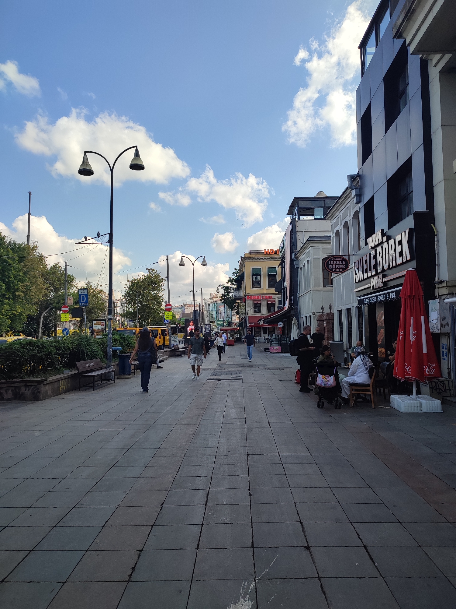 The image depicts a street scene in Kadıköy, Turkey, on a clear day with scattered clouds. A wide, paved sidewalk occupies the foreground, stretching into the distance. Several people are walking on the sidewalk, some singly and some in small groups. On the left side of the sidewalk, a row of benches and lush green bushes line the area, separating the pedestrian walkway from a street with vehicles. Traffic lights and street signs are visible. On the right side of the sidewalk, a commercial building with a prominent sign that reads "Tarihi İskele Börek" dominates the scene. Several umbrellas, one red and branded with what appears to be Coca-Cola, are set up outside the establishment, and a few patrons are seated at tables. Further down the street, more buildings and shops line the sidewalk. The overall impression is of a bustling urban environment.