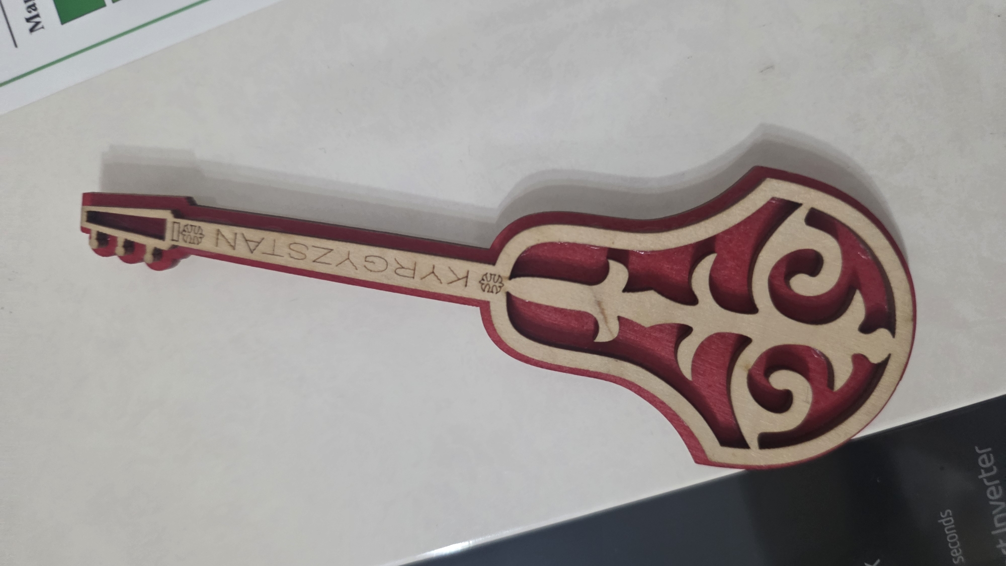 A close-up, top-down shot shows a wooden souvenir shaped like a traditional Kyrgyz musical instrument, possibly a komuz, placed on a white surface. The souvenir is primarily red with intricate, light-colored wood inlays that form decorative patterns. The word "KYRGYZSTAN" is carved vertically into the neck of the instrument. The background is out of focus but appears to be a flat surface, possibly a table or countertop, with a hint of a printed document visible on the right edge of the frame. The lighting is soft and diffused, casting minimal shadows. There are no people or animals present in the image. The souvenir is the sole focal point of the image. The specific location within the city of Chuy, Kyrgyzstan, cannot be determined from the image alone.