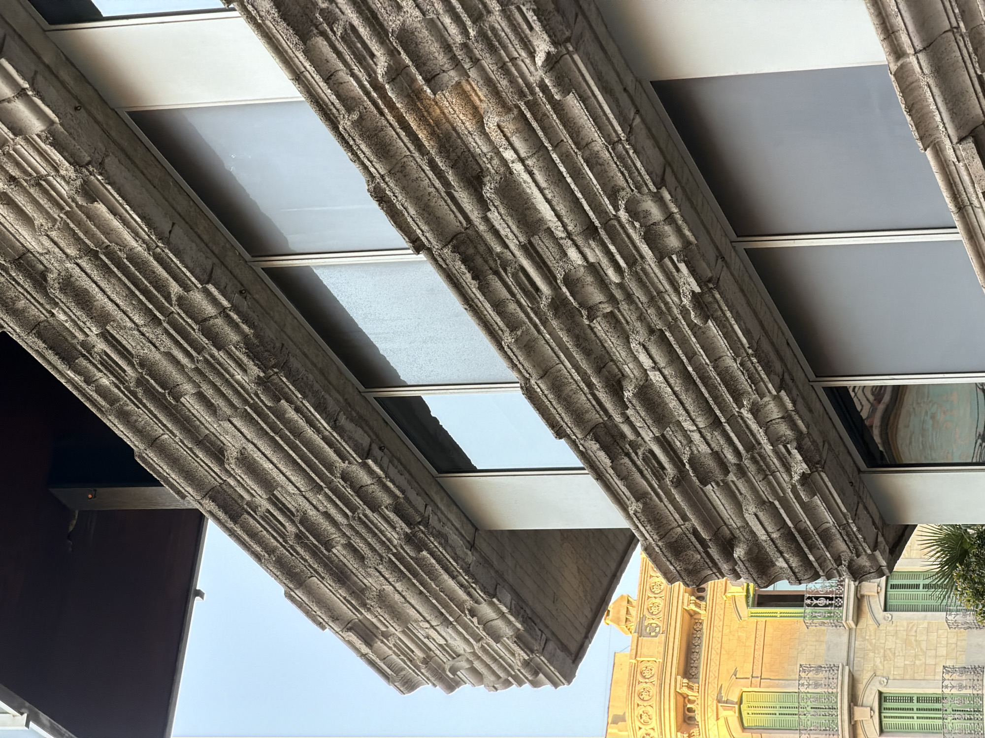 This image captures a close-up perspective of the facade of a building in Barcelona, Spain, featuring a textured, Brutalist-inspired concrete overhang with repetitive horizontal grooving. The overhang casts a shadow on the lower portion of the building. Several windows with reflective, gray panes are visible behind the concrete structure. In the background, a yellow building with green shutters and balconies is illuminated by warm sunlight, indicating it is likely late afternoon or early morning. The sky is a clear, pale blue. The overall scene depicts an architectural detail of a modern building juxtaposed with a more traditional building in its vicinity.