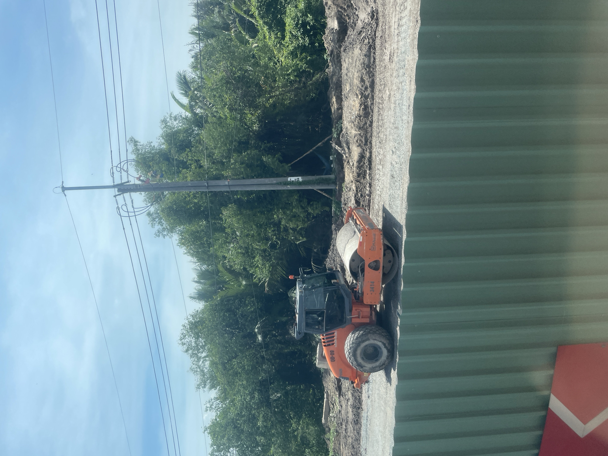 This image captures an outdoor scene with a construction site in Dong Nai City, Vietnam. In the foreground, a green corrugated metal fence runs horizontally across the bottom of the frame. Behind the fence, an orange road roller is positioned on a gravel surface. The road roller is a piece of heavy machinery with a large cylindrical drum at the front and tires at the back. It has the markings "QD-09" and "3414" visible on its body, along with a "HAMM" logo. The background features dense green foliage, including trees and palm fronds, suggesting a natural or semi-rural environment. A utility pole with wires stretches upwards into a partly cloudy blue sky. The ground around the road roller appears to be dirt and construction debris, indicating ongoing work.