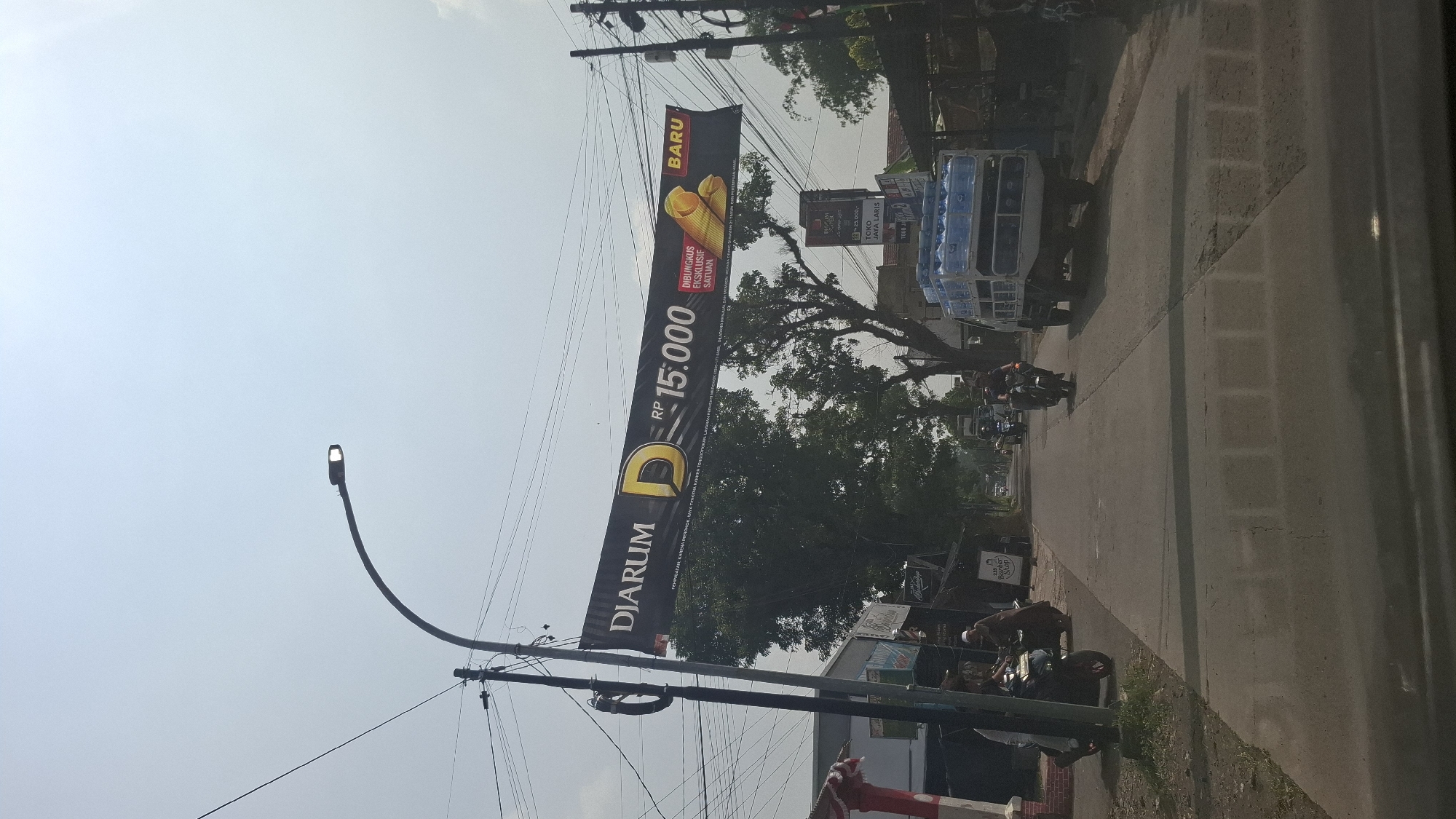 A street scene in Sukasari, Indonesia, features a black banner with yellow lettering reading "DJARUM" and "RP 15.000" stretched across the street. A delivery truck loaded with bottled water is visible, as are several motorcycles and pedestrians. The environment is outdoors with a clear sky and trees lining the street.
