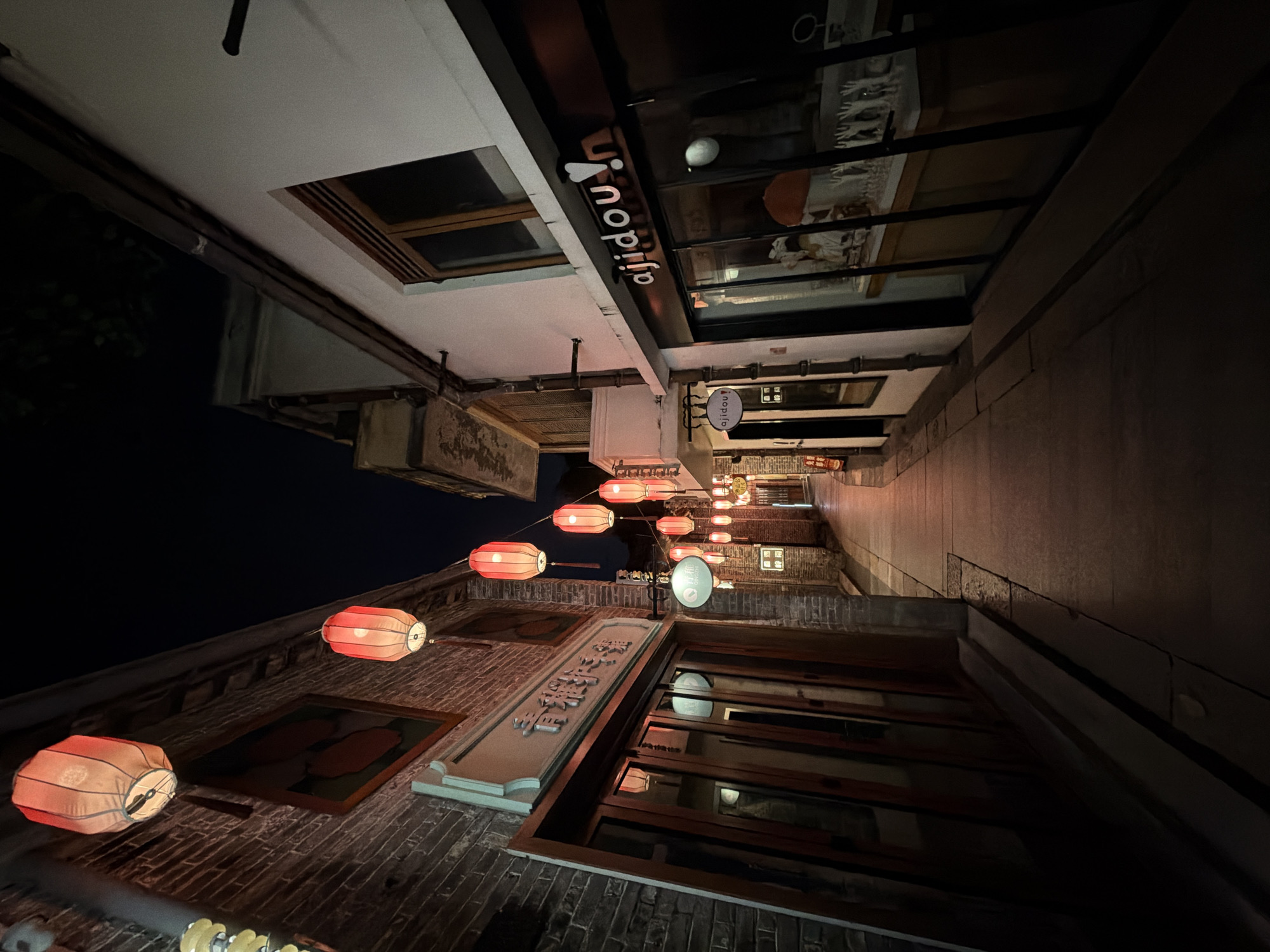 The image captures a narrow street at night, illuminated by numerous red lanterns strung across the pathway. The architecture suggests a historic district, with buildings on either side of the street constructed of brick and white plaster. The street itself is paved with stone. On the left, a brick building features a wooden-fronted shop with glass doors reflecting the lanterns. A sign with Chinese characters is mounted above the entrance. On the right, a white building has large display windows showcasing merchandise and a prominent sign reading "ajidou!". The scene is devoid of people, creating a quiet and atmospheric ambiance. The overall impression is one of a charming, well-lit alleyway in an urban setting, likely a commercial or cultural area. The presence of Chinese characters on signage indicates a location within China. Considering the provided context that the image is from Chancheng District, China, it likely depicts a scene from an older part of the city, possibly a pedestrian street or shopping area that is decorated with traditional lanterns.