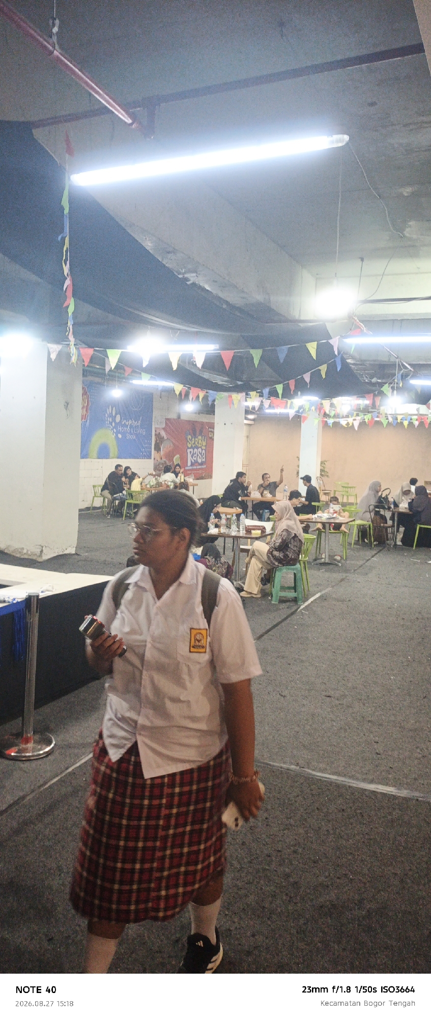 The image depicts an indoor market or food court setting. A young person wearing a white collared shirt, a red and yellow plaid skirt, white socks, and black sneakers is the primary subject in the foreground, walking and holding a device that resembles a flashlight. The person is wearing glasses and has dark hair pulled back. Behind them, a crowd of people is seated at tables, some eating or conversing. The environment is illuminated by fluorescent lights and decorated with colorful triangular flags strung across the ceiling. In the background, banners with text are visible, including one that appears to advertise a "Home & Living Show" and another with the text "SerBu Rasa". The lower portion of the image contains metadata indicating the photo was taken on August 27, 2026, at 15:18, with a camera setting of 23mm f/1.8 1/50s ISO3664. The location is identified as Kecamatan Bogor Tengah, Bogor, Indonesia.