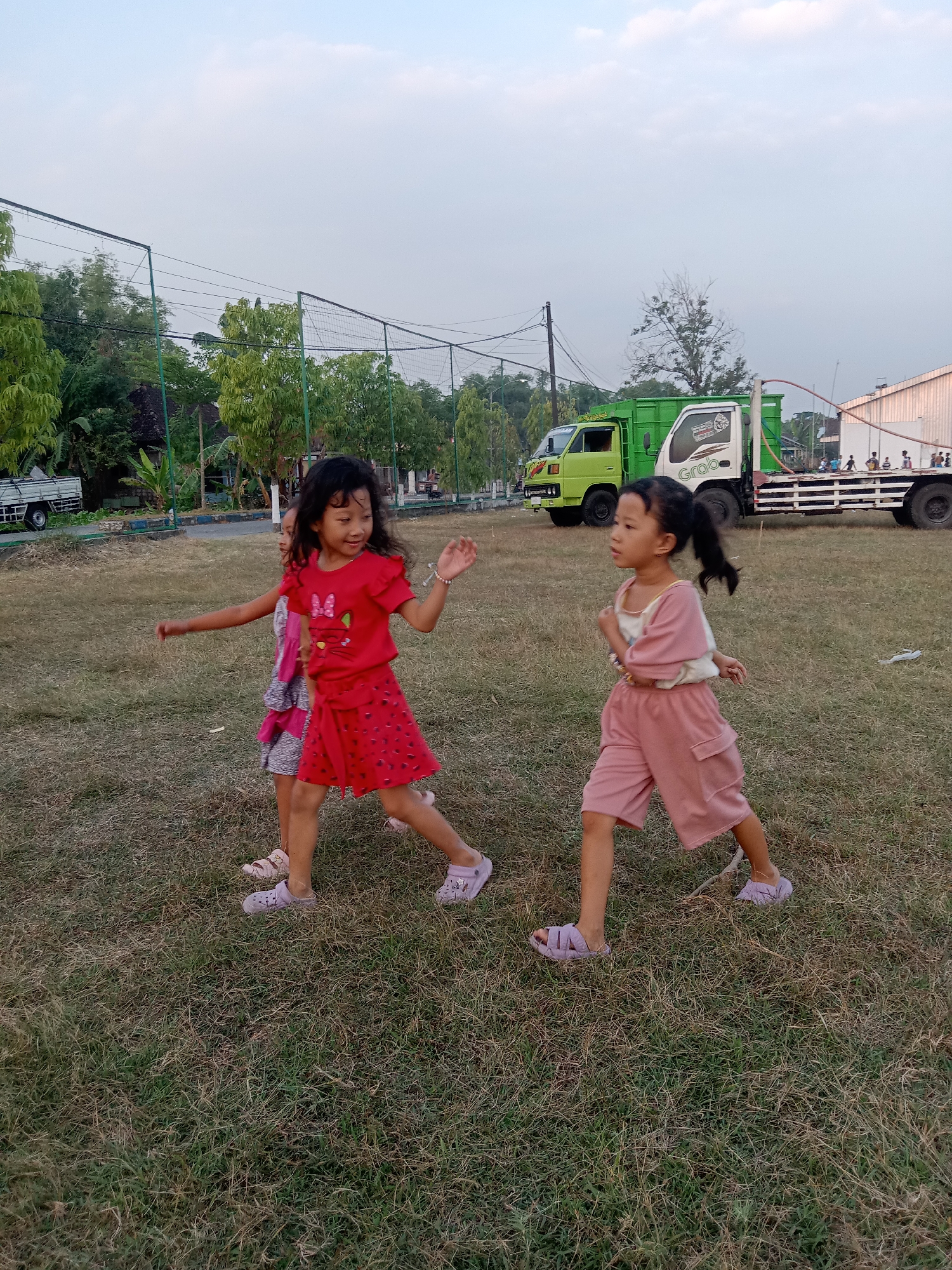 Three young girls are running on a grassy field in Stiaji, Indonesia, under a clear sky. The girl in the foreground, on the right, wears a pink top and shorts, with her dark hair in a ponytail. To her left, a girl in a red dress with a heart pattern runs with her arm outstretched. Further to the left, partially obscured, is a third girl in a pink and patterned dress. In the background, two trucks are parked on a road, one green and one white with "Grab" branding. Trees and a fence line the perimeter of the field.