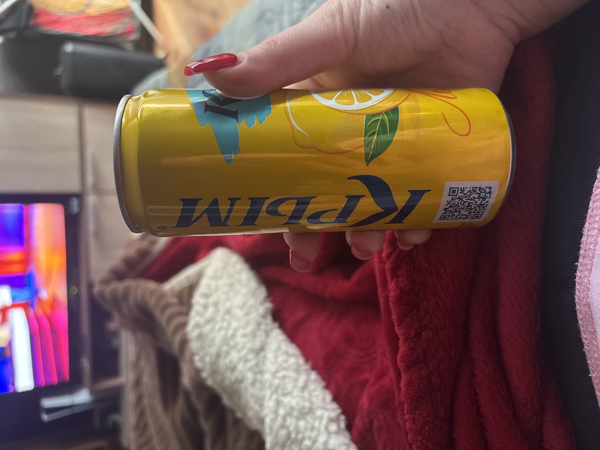 A person's hand with red nails is holding a yellow can of KPIM lemon-flavored drink. The can has an image of a lemon and a QR code. The text "KPIM" is written in dark blue, but it is reversed in the image. The background includes a television screen with colorful images and a red and white blanket. The image does not contain sufficient information to ascertain that the image was taken in Chornomorskoe, Ukraine.