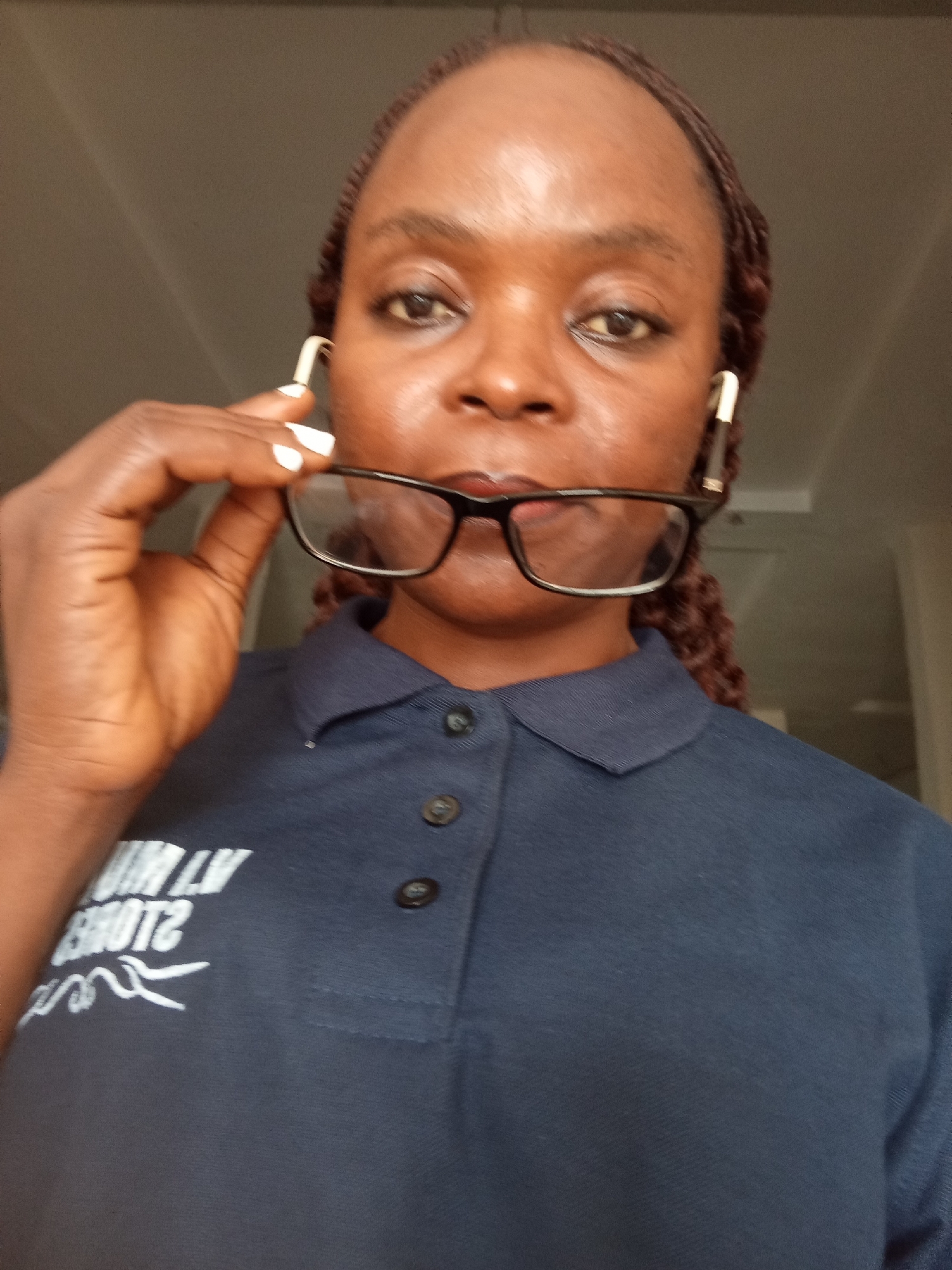 A close-up, eye-level, medium shot shows a Black woman with braided hair and glasses resting on her nose. She is holding the frames of her black-rimmed glasses with her right hand, which is positioned near her face. The woman is wearing a navy blue polo shirt with white text and a graphic design on the left side of her chest. The text appears to be "LUVM2 STORES" in a reversed orientation. The background is a neutral, blurry gray. The lighting is soft, with light coming from the front, illuminating the woman's face.