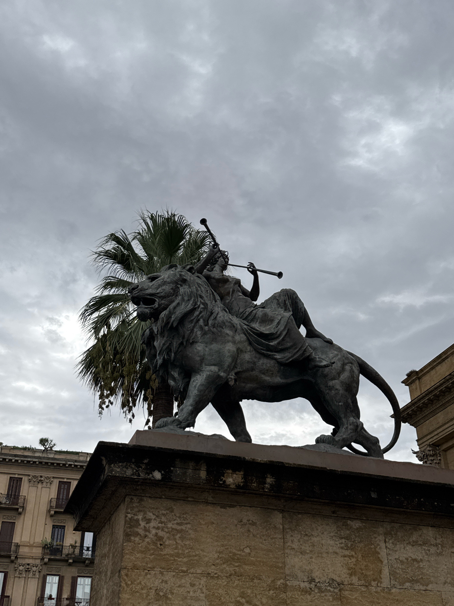 A bronze statue of a woman riding a lion is positioned on a stone pedestal in Palermo, Italy. The woman is seated on the lion's back, holding a horn to her lips. Behind the statue, a palm tree with green fronds is visible, its fronds reaching upwards. The background is dominated by a cloudy, overcast sky. To the left and right of the statue, parts of buildings with arched windows and balconies can be seen. The statue is located in front of the Teatro Massimo, one of the largest opera houses in Italy.