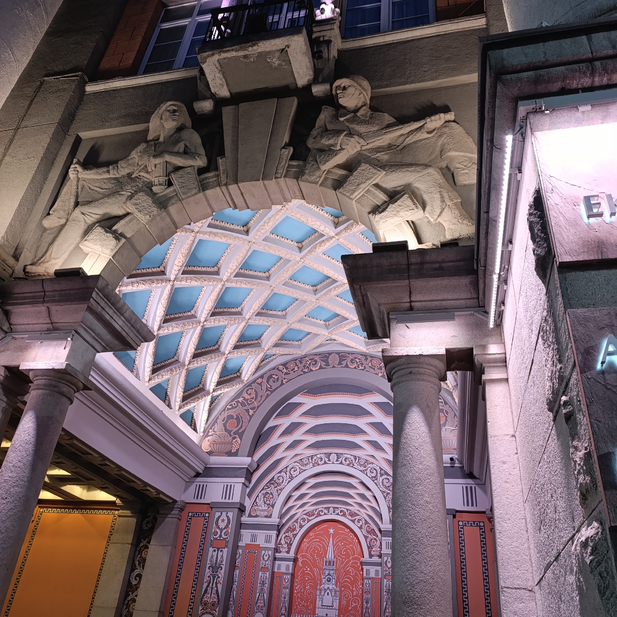 This is an image of the entrance to the Main Post Office in Yekaterinburg, Russia. The building's facade features two sculptures above the archway: one depicting a woman holding a rolled-up document, and another depicting a man writing. The archway leads into an interior space with a blue coffered ceiling. The walls are decorated with a mural depicting a stylized cityscape and elaborate decorative patterns. Two columns flank the entrance. The lighting suggests it is nighttime.