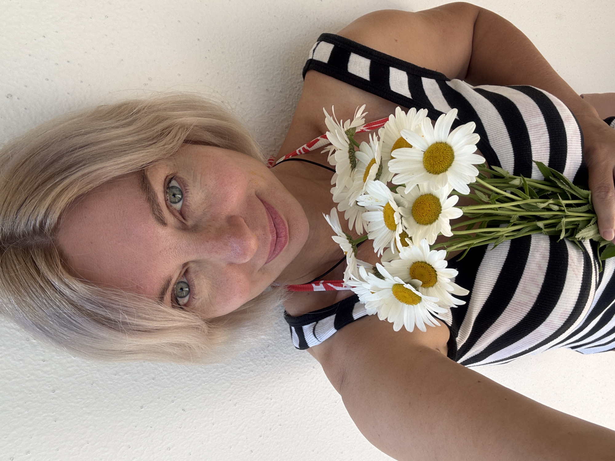 A woman with short, blonde hair and blue-green eyes is holding a bouquet of daisies in front of her chest. She is wearing a black and white striped tank top with red and white accents on the straps. The background is a textured white wall. The scene appears to be outdoors, possibly in the city of Sirius, Russia, given the context. The woman is looking directly at the camera with a slight smile.