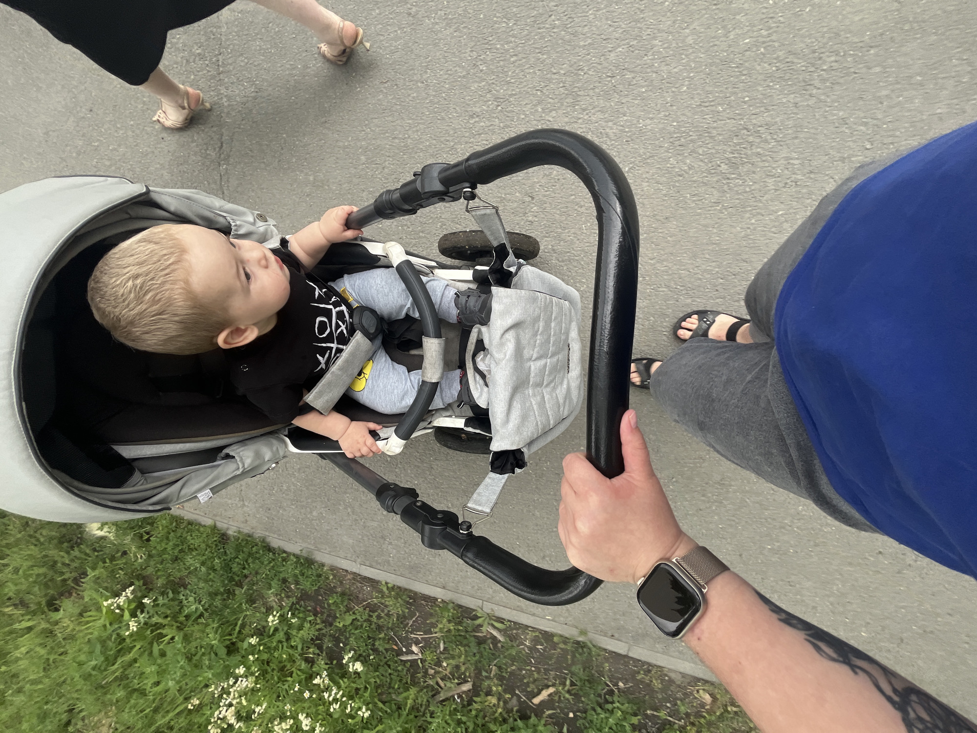 An individual's arm and hand, wearing a smartwatch and a tattoo, are visible at the bottom left of the frame, pushing a stroller. The stroller contains a young child with blonde hair, wearing a black t-shirt and grey pants, seated and looking to the right. To the right of the stroller, a person's legs are visible, wearing sandals and a dark dress or skirt, walking away from the camera. The scene takes place outdoors on a paved path, with green grass and small white flowers lining the left side of the path. The image appears to be taken from a first-person perspective, looking down at the stroller and the path ahead.