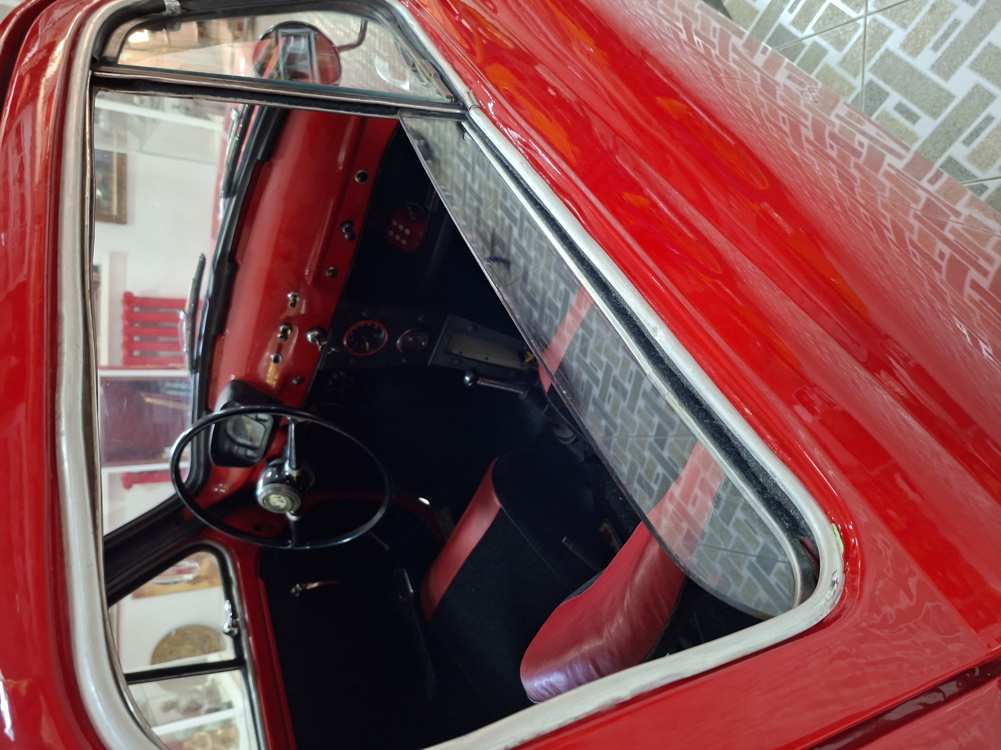 The image shows the interior of a red vintage car, viewed through the open driver's side window. The dashboard is red with black accents and a black steering wheel. A portion of the driver's seat, upholstered in red and black, is visible. The floor is tiled with a geometric pattern. The car is parked indoors, possibly in a garage or showroom, with what appears to be an exhibition space visible through the car's windows, including some artwork and display items. The context suggests this might be a museum or a private collection in Ialoveni, Moldova, given the image's origin. There are no people in the image.