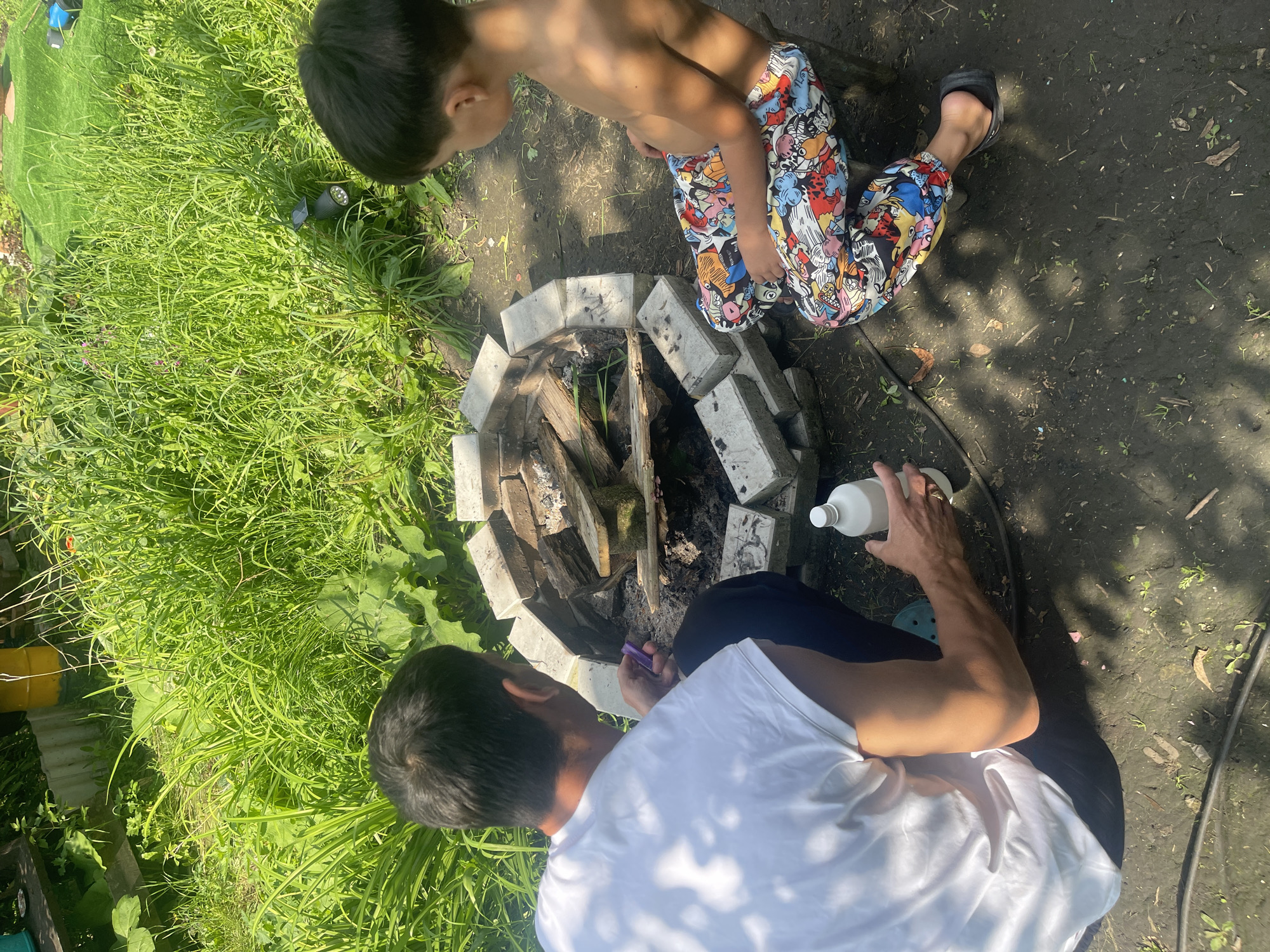 The image depicts two individuals, an adult male and a child, engaged in preparing a fire pit in an outdoor setting. The adult, wearing a white t-shirt and black pants, is positioned on the left, holding a purple object and a white bottle. The child, shirtless and wearing colorful patterned pants, sits on the right, facing the fire pit. The fire pit, constructed from bricks, contains wood. The environment is characterized by lush green grass and foliage, suggesting a backyard or park area. The ground is predominantly dark soil with some scattered debris. The lighting indicates it is daytime, with sunlight casting shadows. The location is identified as Barnaul, Russia.