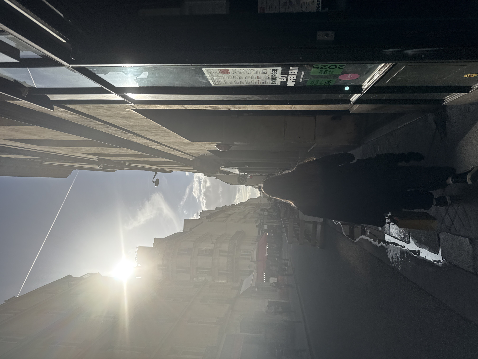 The image portrays a narrow, sunlit street in Paris, seen from a slightly elevated perspective. The primary subject is a person, silhouetted by the bright light, walking away from the viewer towards the end of the street. They have light-colored hair and are wearing a dark coat and dark footwear with white details.
The street is flanked by multi-story buildings, typical of Parisian architecture, featuring multiple windows and some balconies. The ground appears to be a mix of pavement and potentially wet areas or puddles, reflecting some of the light.
On the right side of the frame, a dark storefront is visible, featuring large windows and signage. Visible text on this sign includes "BIG BURGER" and "EAT DIFFERENT," indicating a restaurant, likely a burger establishment. Other legible text includes "2025," "Uber Eats," and "Doordash," along with the Deliveroo icon, suggesting food delivery services are offered. A menu or list of items is also partially visible on the sign.
The time of day is bright and sunny, with strong backlighting from the left side of the frame, creating significant lens flare and long shadows. The sky is clear with some scattered clouds and a visible contrail from an airplane. The overall atmosphere is that of a bright, everyday urban scene.