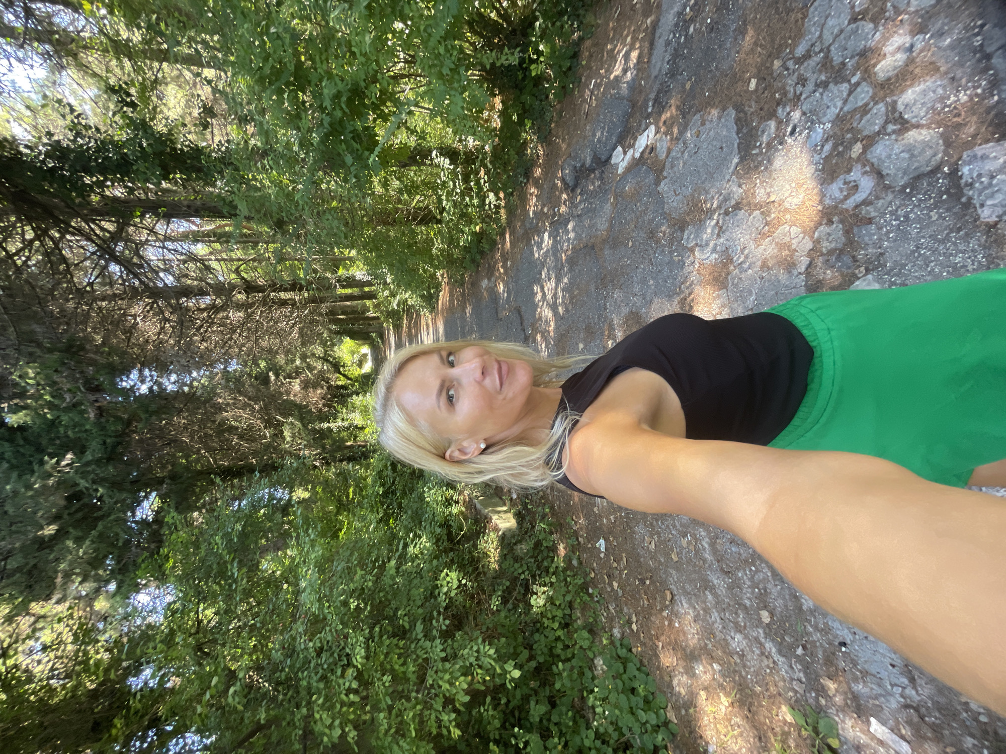 A blonde woman with fair skin, wearing a black sleeveless top and a bright green skirt, is taking a selfie on a sun-dappled, unpaved path. The path is lined with dense, lush green trees and foliage, characteristic of a park or forest area. The woman's right arm is extended towards the camera, holding the device. She is looking towards the camera with a slight smile. The path appears to be a mix of dirt, gravel, and cracked asphalt, with fallen leaves scattered on the surface. The trees are tall and slender, with some appearing to be cypresses, and others covered in ivy. The lighting suggests it is daytime, with sunlight filtering through the canopy, creating patches of light and shadow on the path. The environment appears natural and somewhat wild, suggesting a location like the Dendrarium Botanical Garden in Sochi, Russia, known for its diverse flora and walking paths.